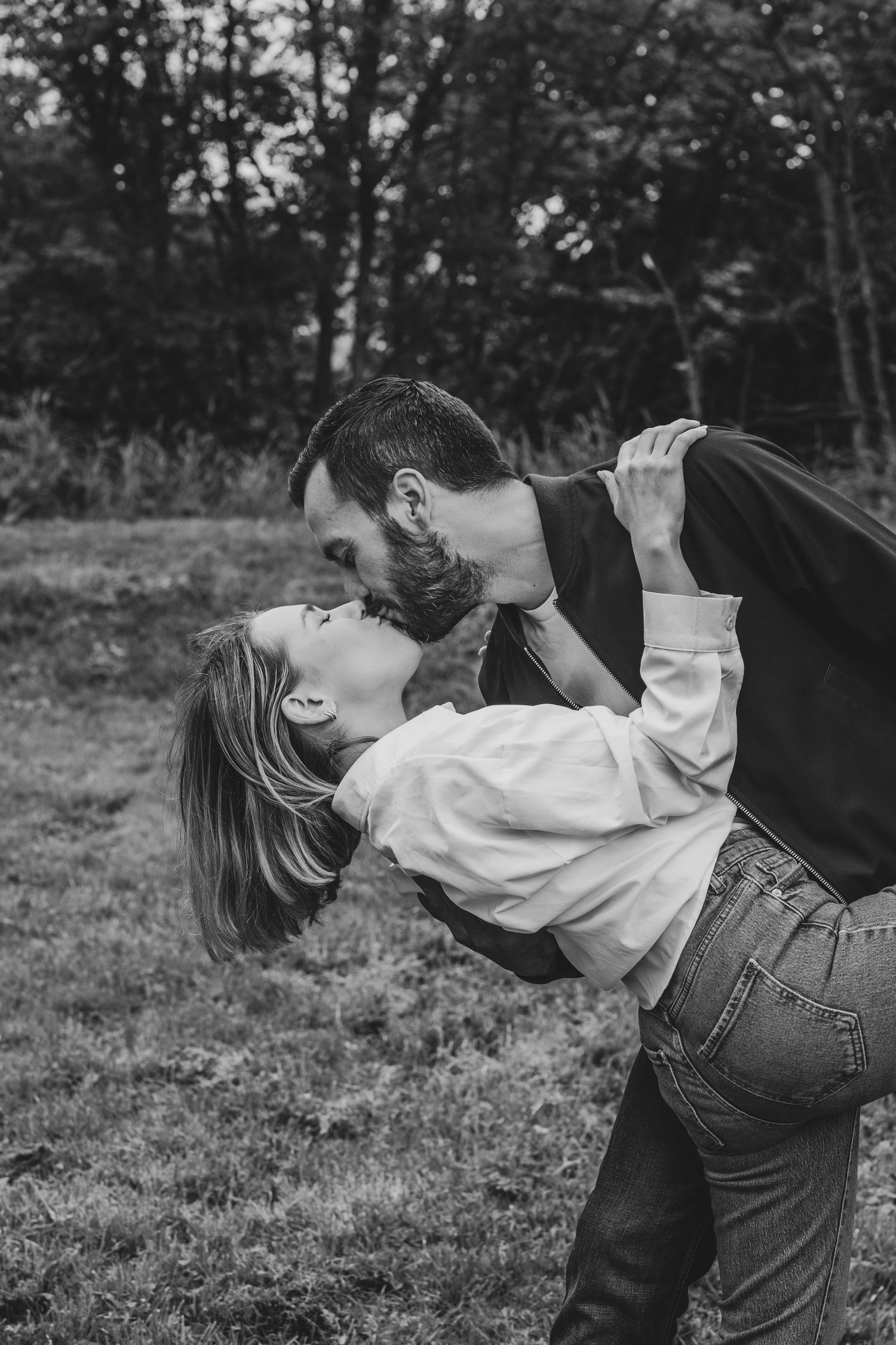 Black and white photo of a romantic couple sharing a passionate dip kiss in a lush outdoor setting.