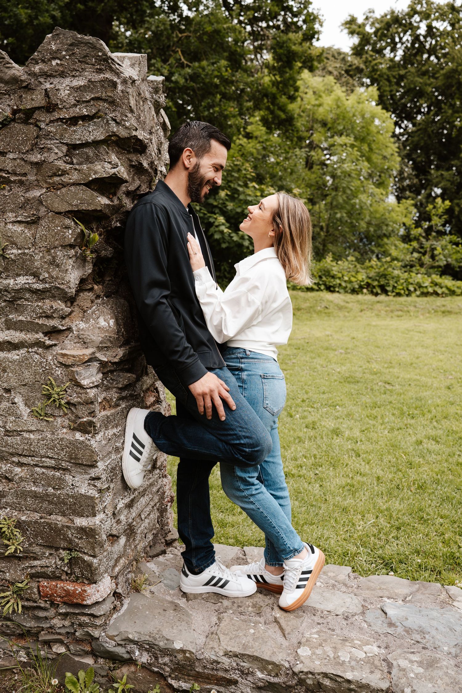 A couple laughing and embracing against a rustic stone wall in a lush green park, wearing casual jeans and sneakers.