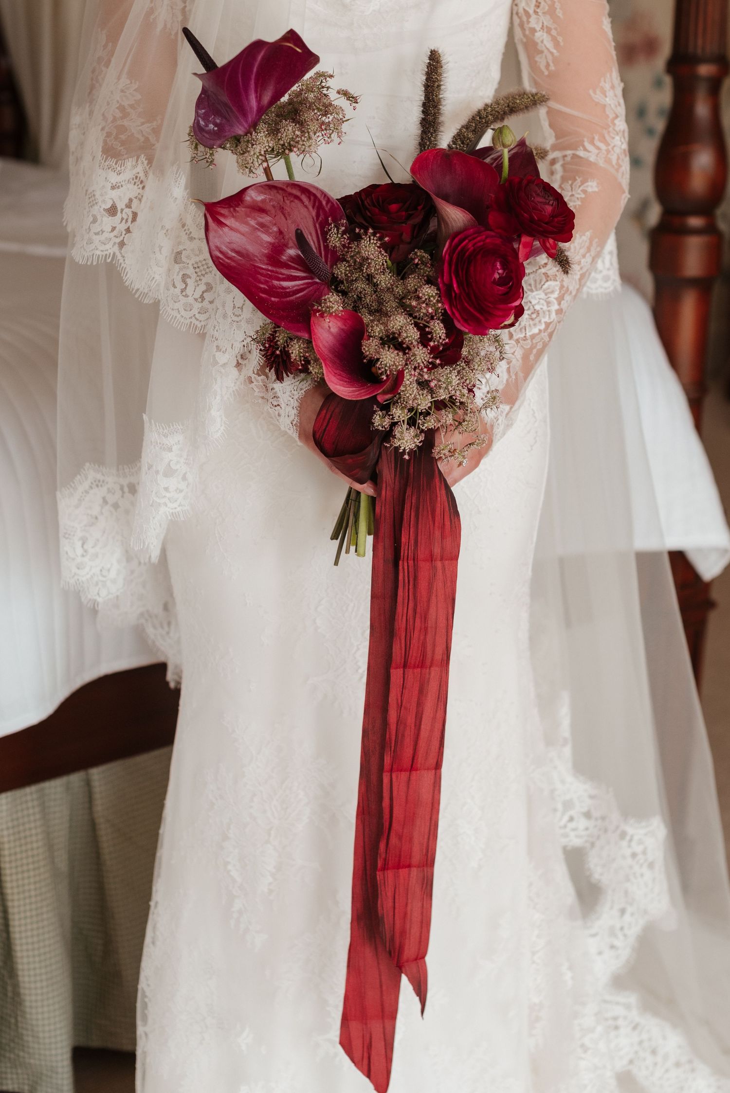 Bride holding deep red bridal bouquet of roses and anthuriums with long ribbon against lace wedding gown.