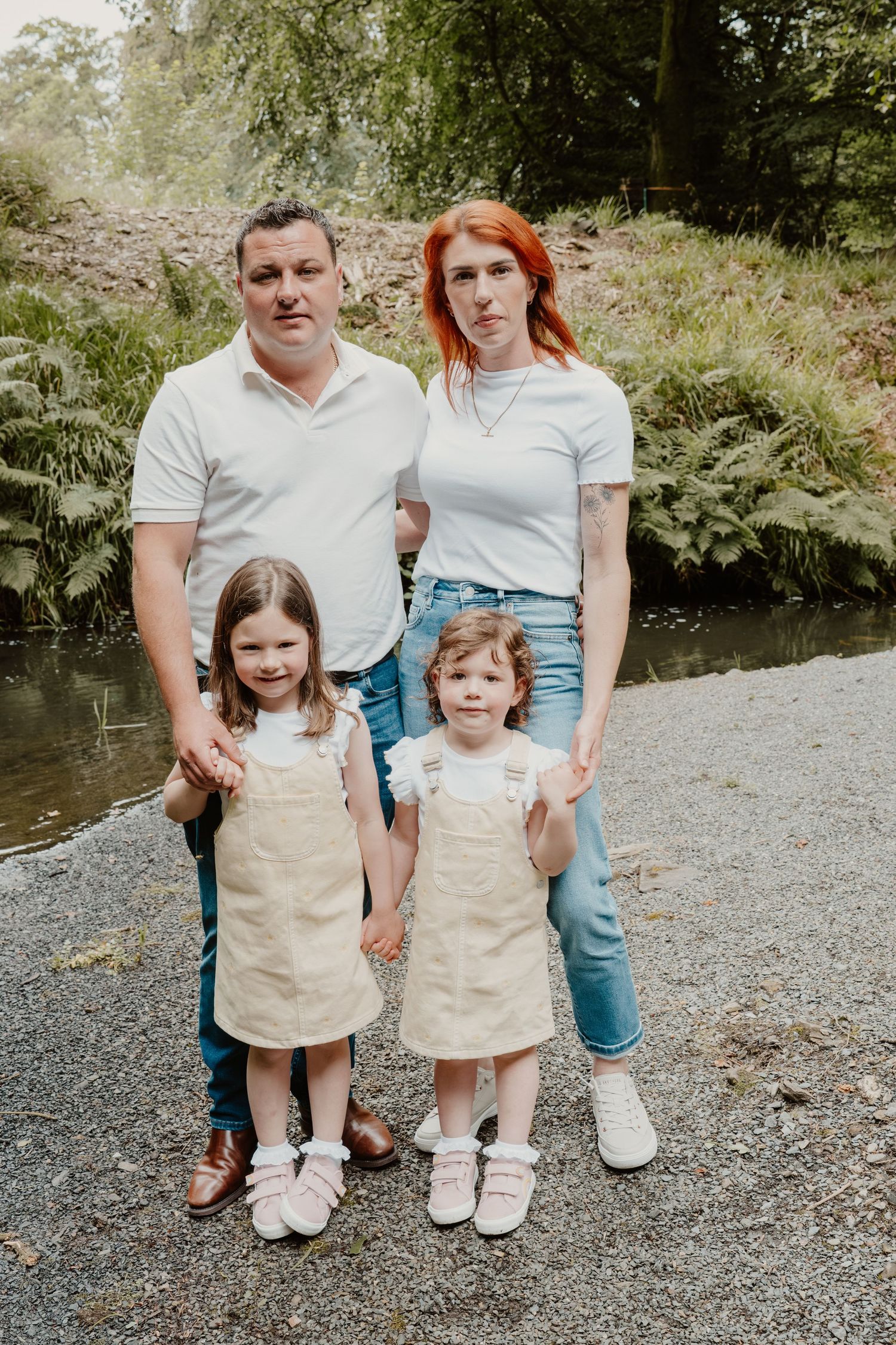 Family of four posing outdoors near a stream, parents in white tops with two young girls in matching beige pinafore dresses.