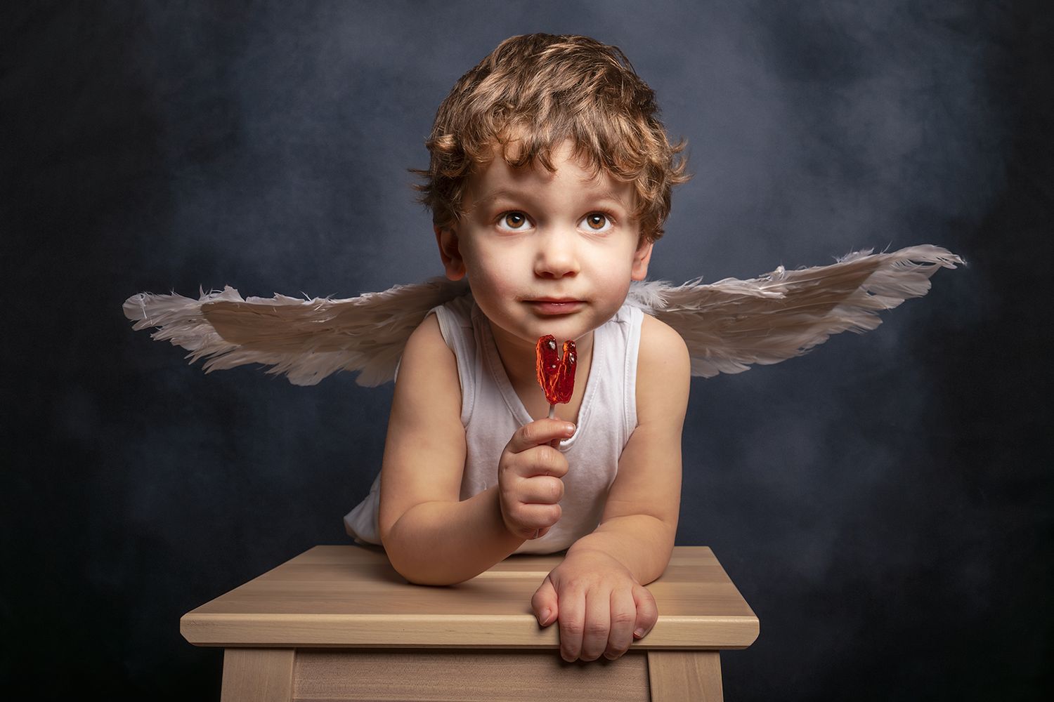 Adorable young boy dressed as Cupid with angel wings holding a red lollipop, posing on a wooden stool.