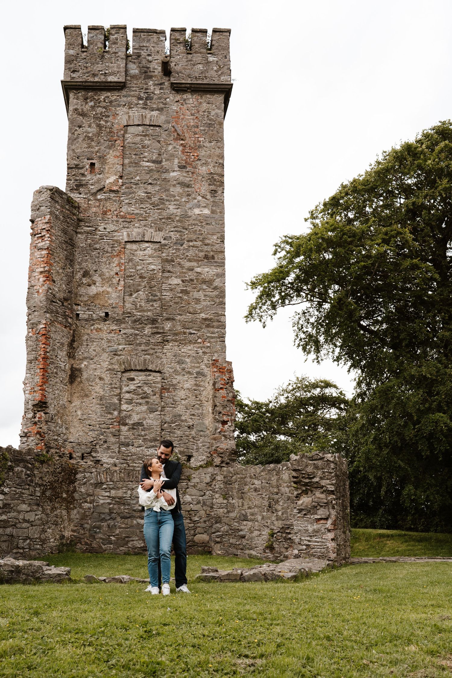 A couple embraces on green grass before a historic stone castle tower ruin with battlements and a large tree nearby.
