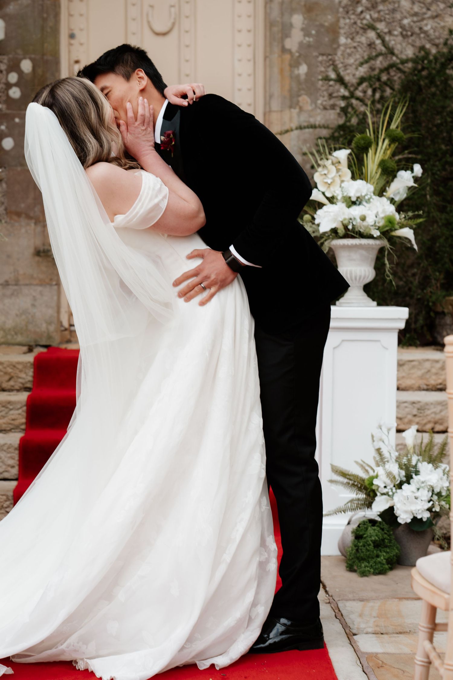 Bride and groom sharing their first kiss on a red carpet aisle, surrounded by white floral arrangements.