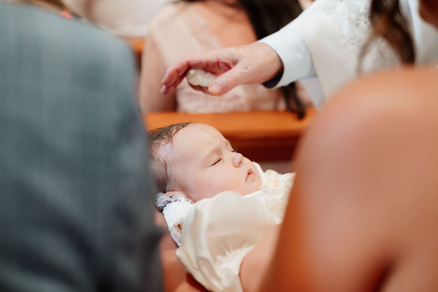 A sleeping baby in white clothing during a baptism ceremony with a hand gently placed on their head.