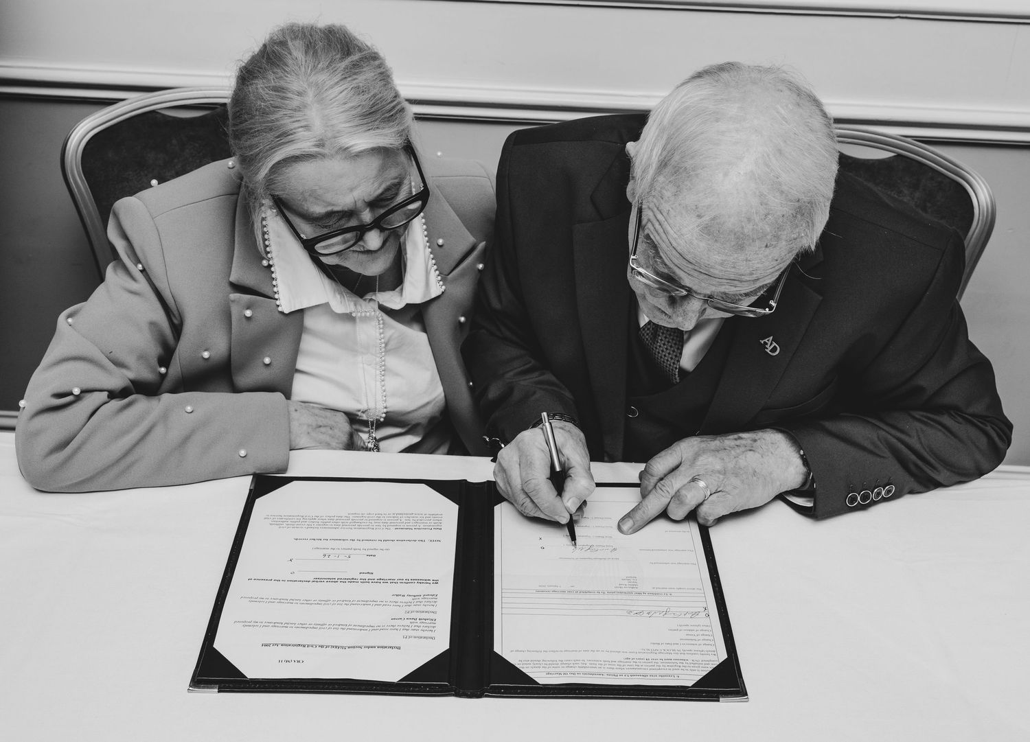 Elderly couple signing official documents together at a table in a black and white photo.