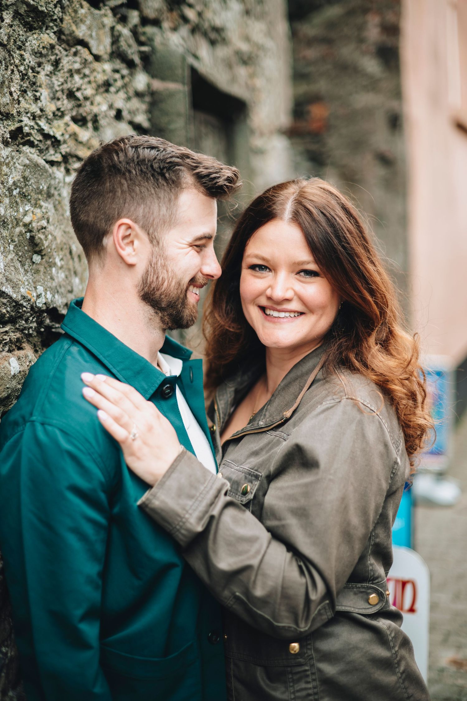 Smiling couple leaning against a stone wall, woman in olive jacket embracing man in teal shirt outdoors.