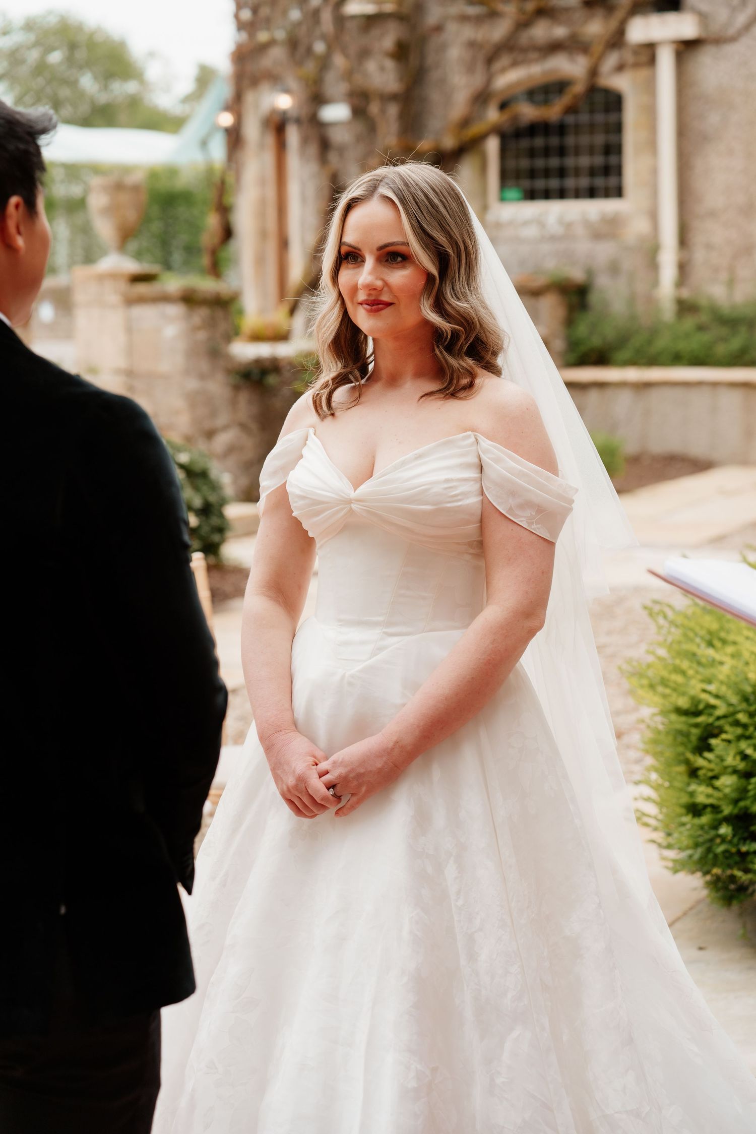 Bride in off-shoulder lace ball gown and veil stands outdoors at stone venue during wedding ceremony.