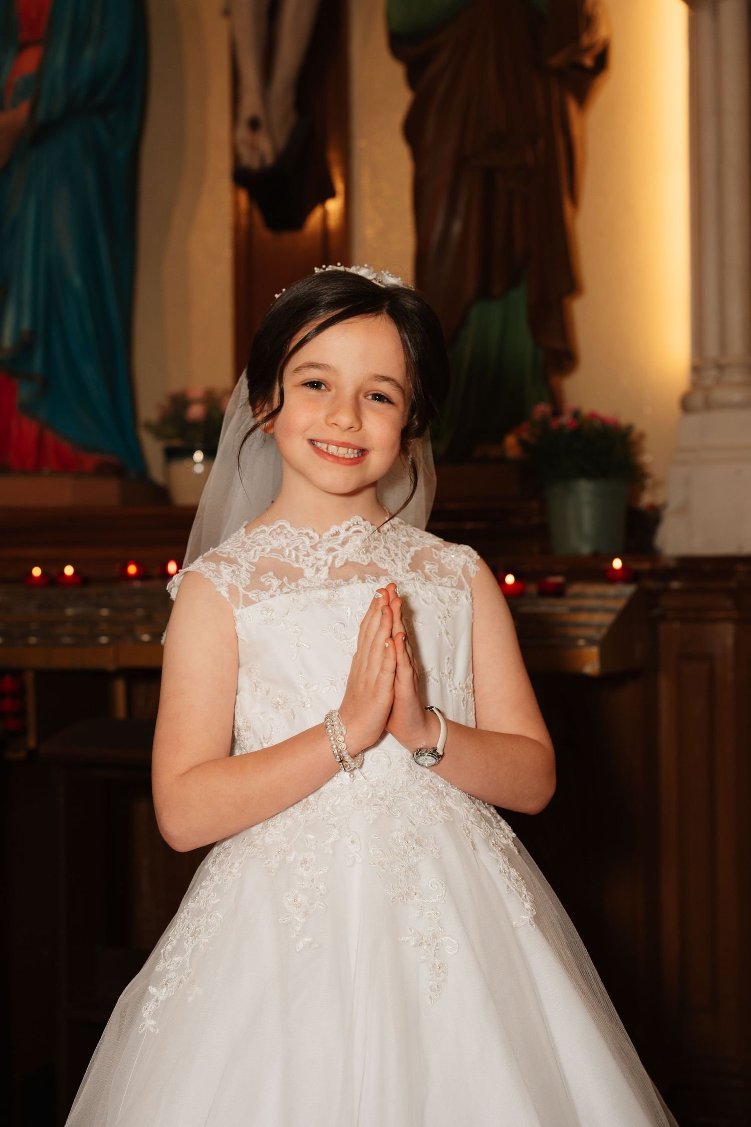 Young girl in white lace First Communion dress and veil, hands clasped in prayer, smiling in a church.