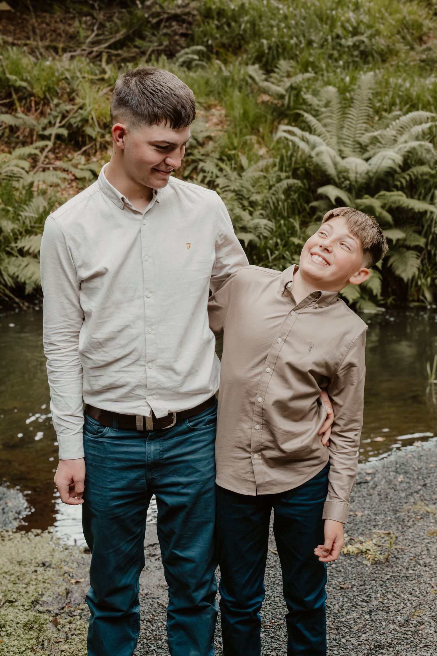 Two smiling young men in button-up shirts stand together outdoors near a stream surrounded by ferns.