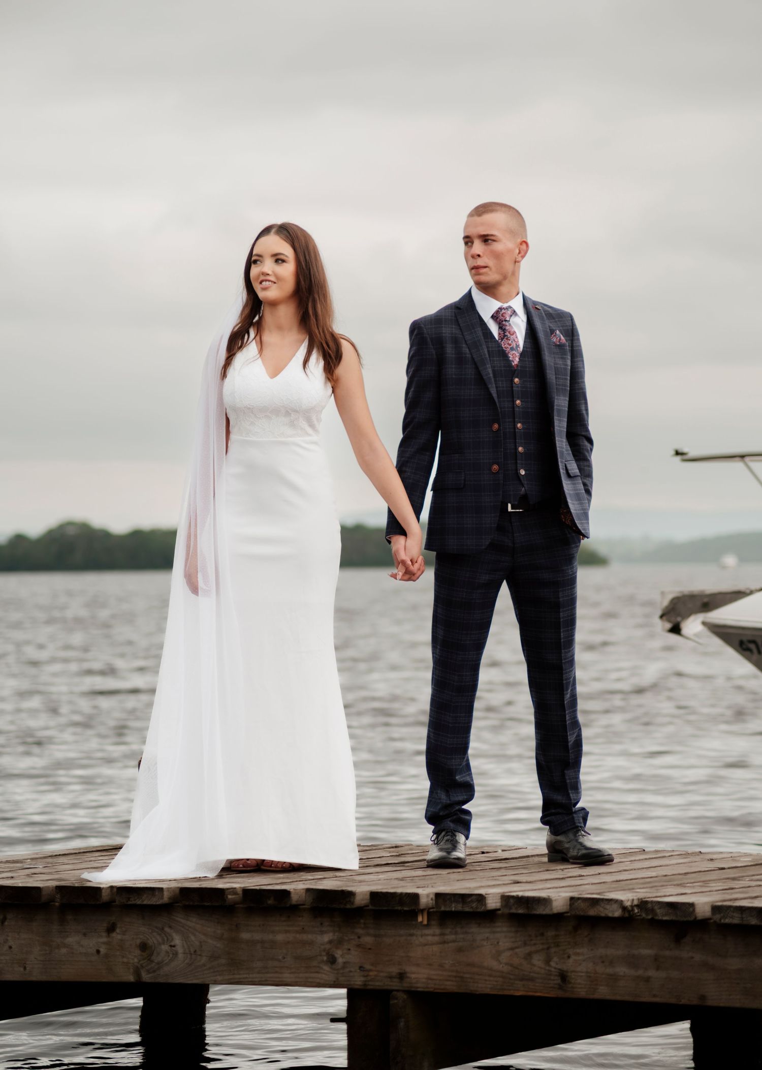 Bride and groom holding hands on a lakeside dock, she in white gown, he in navy plaid suit.