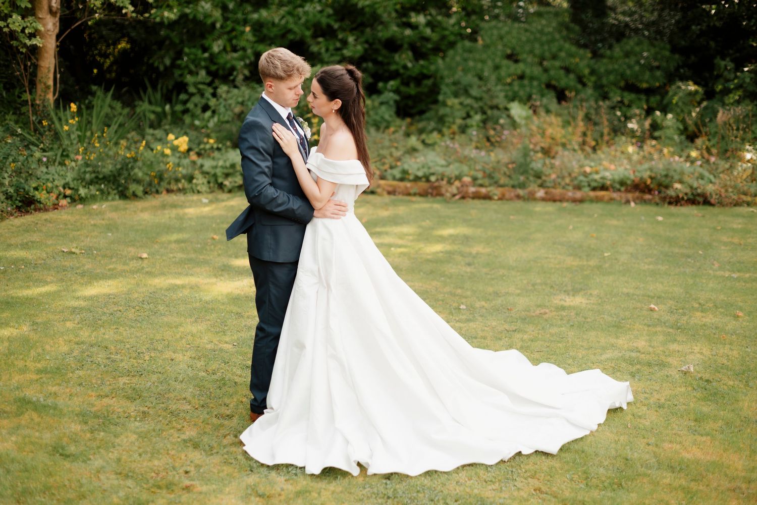 Romantic wedding couple embracing on a lush green lawn surrounded by tall trees in soft light.