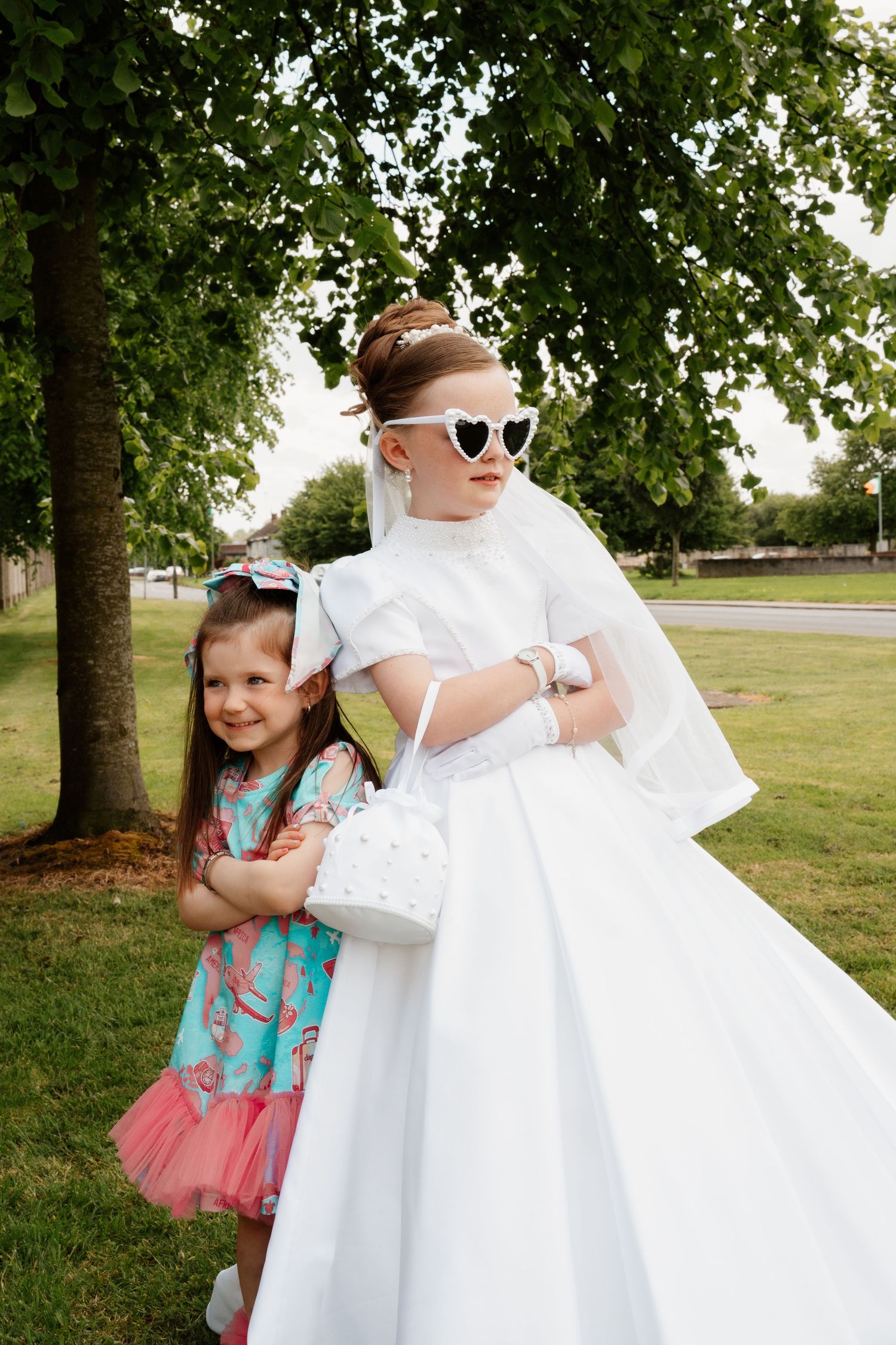 Two young girls in white communion dress with veil and sunglasses posing outdoors with smaller girl in blue floral dress.