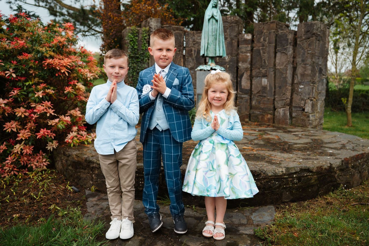 Three children in formal attire pose with hands clasped in prayer near a religious statue in a garden.