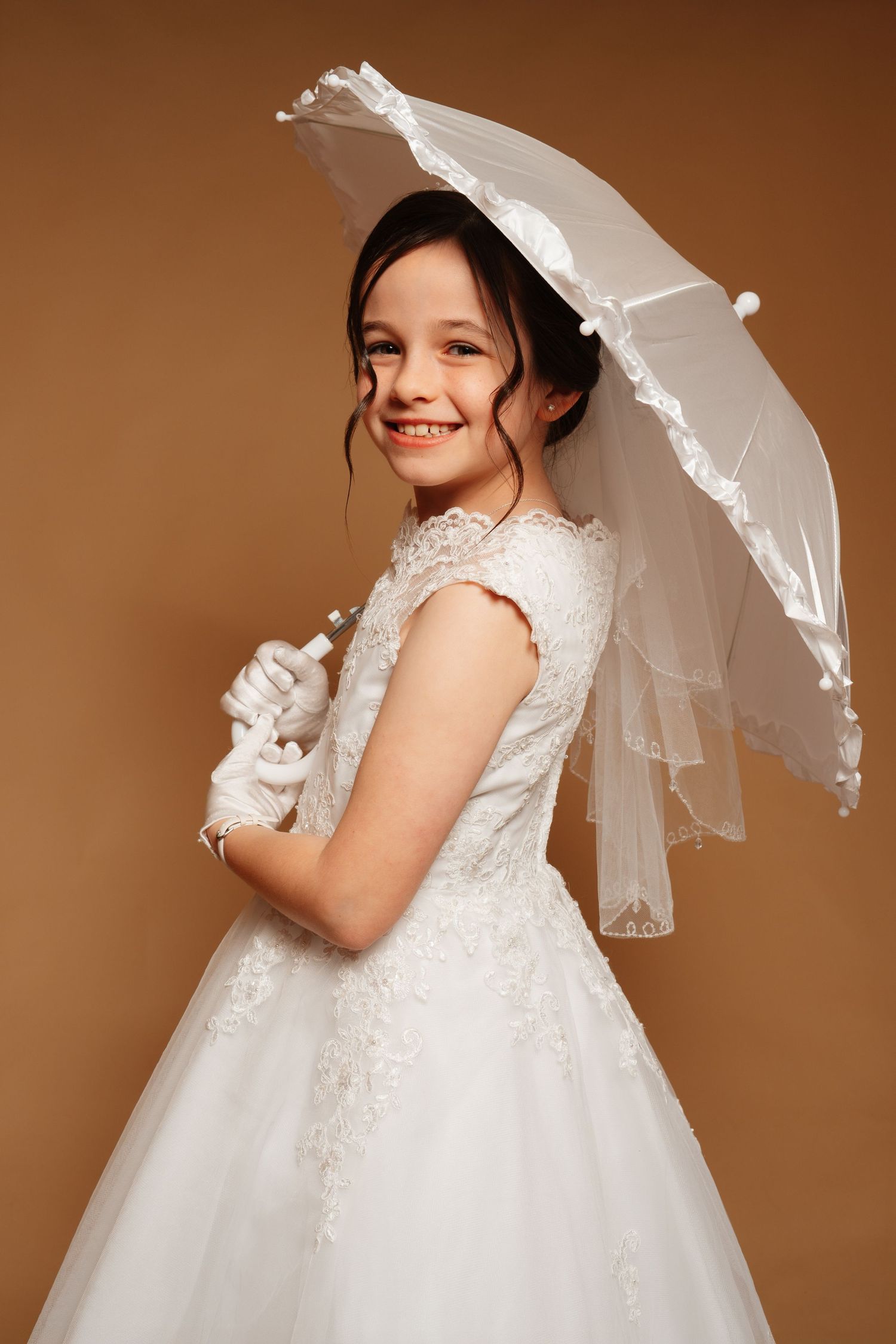 Smiling young girl in white lace dress and gloves holding a white parasol umbrella on beige background.