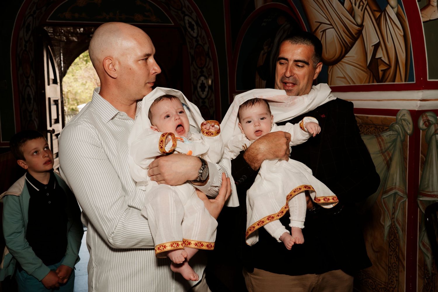 Two men hold twin babies dressed in white christening gowns inside an Orthodox church.