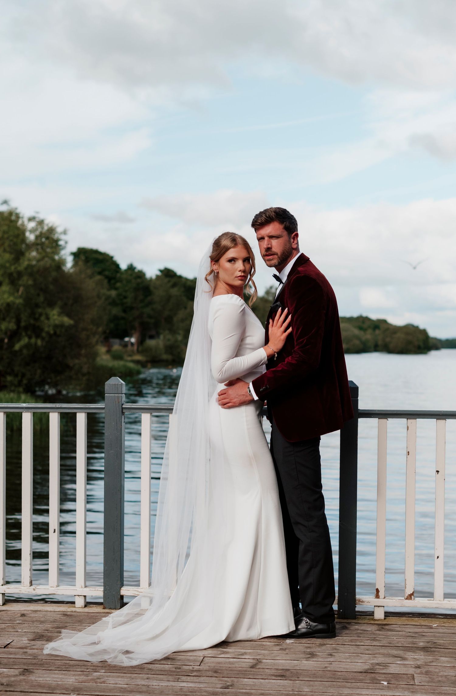 Bride in long-sleeve white gown with cathedral veil embraces groom in burgundy velvet suit on lakeside dock.
