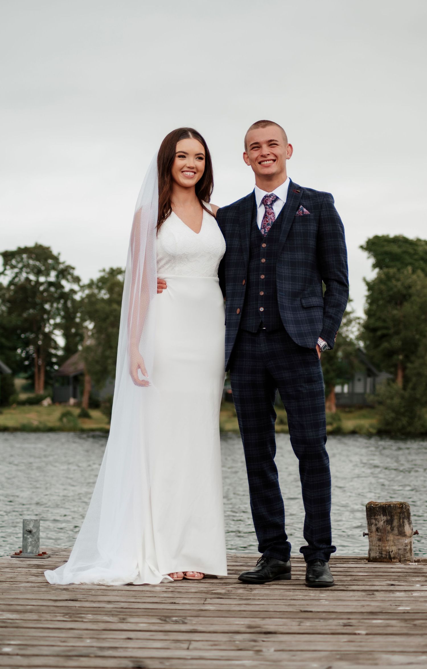 Smiling bride and groom posing on a wooden dock by a lake, bride wearing veil and fitted white gown.