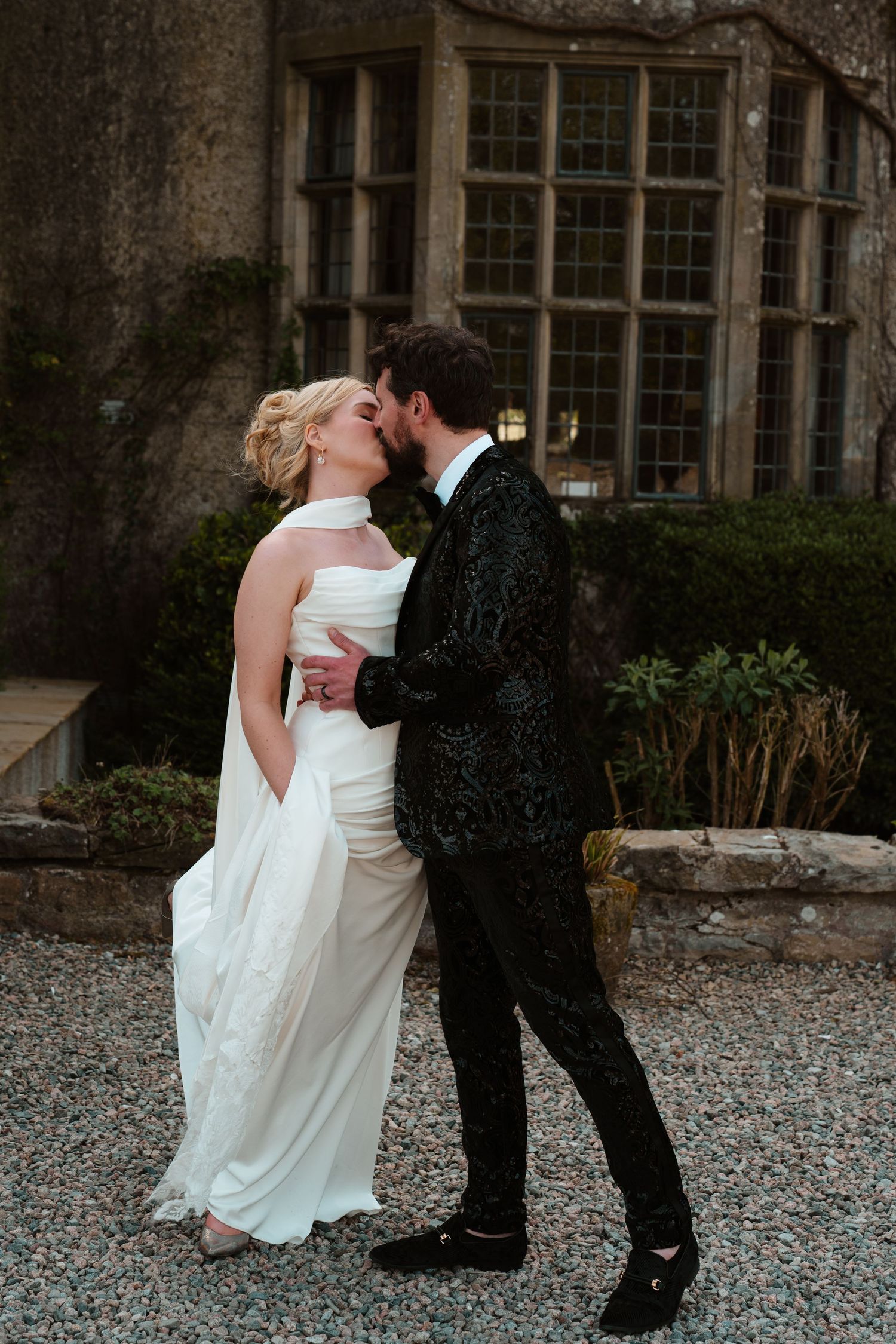 Bride and groom sharing romantic kiss outside historic stone manor in elegant wedding attire.