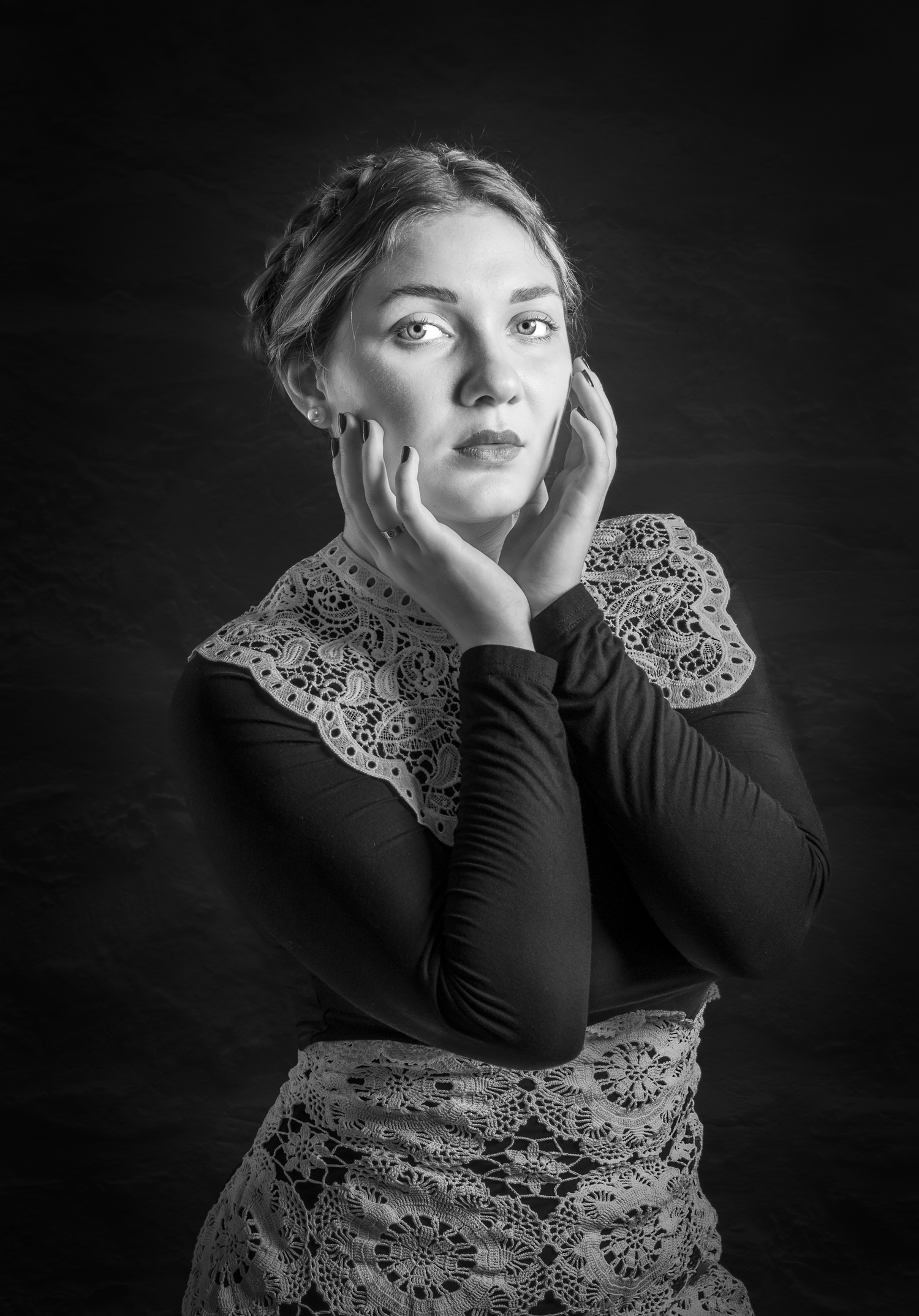 Black and white portrait of a young woman with braided hair, hands framing her face, wearing lace-trimmed outfit.