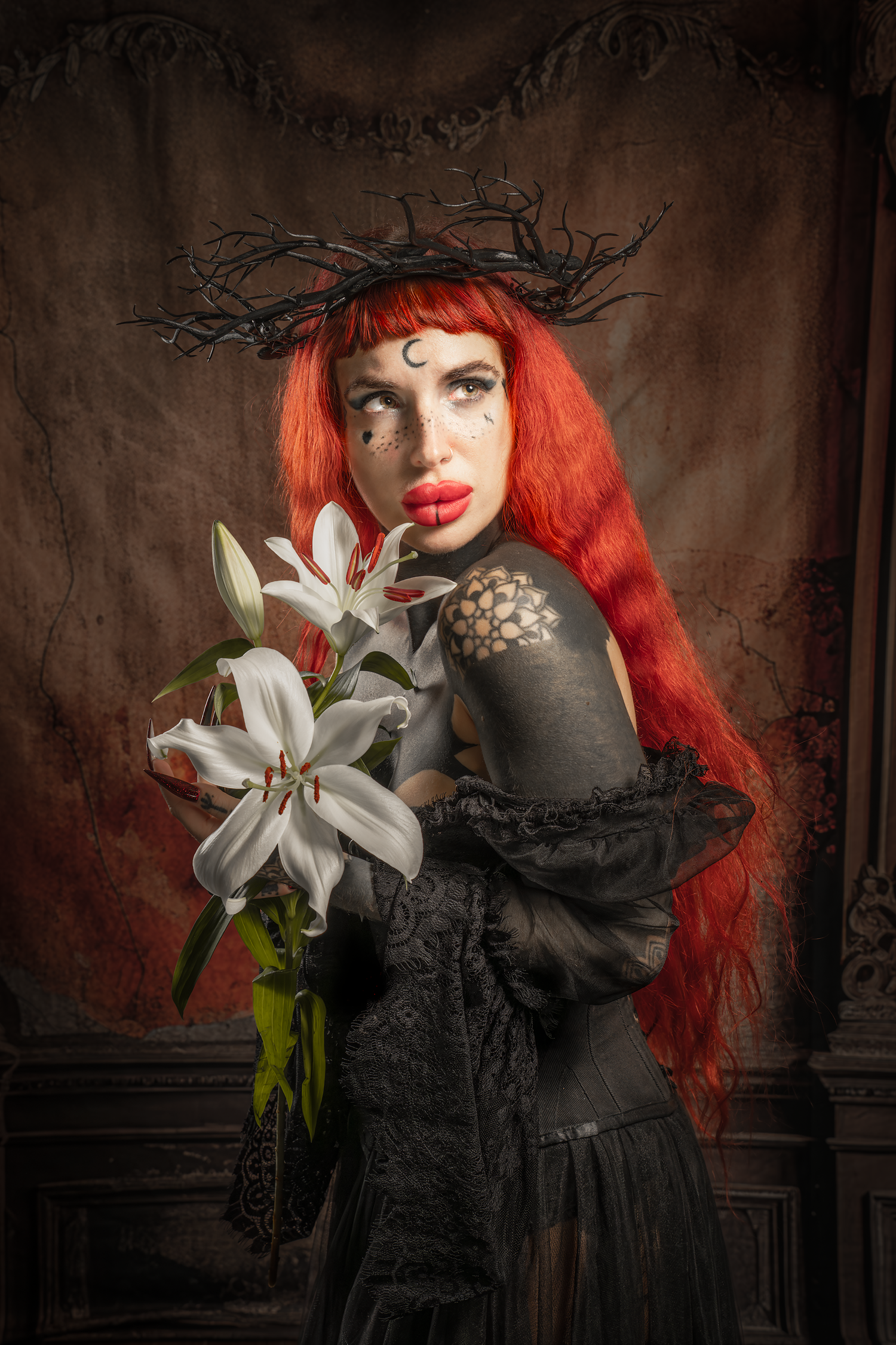 Red-haired woman in gothic black bodypaint holding white lilies, wearing thorn crown, dark dramatic portrait photography.