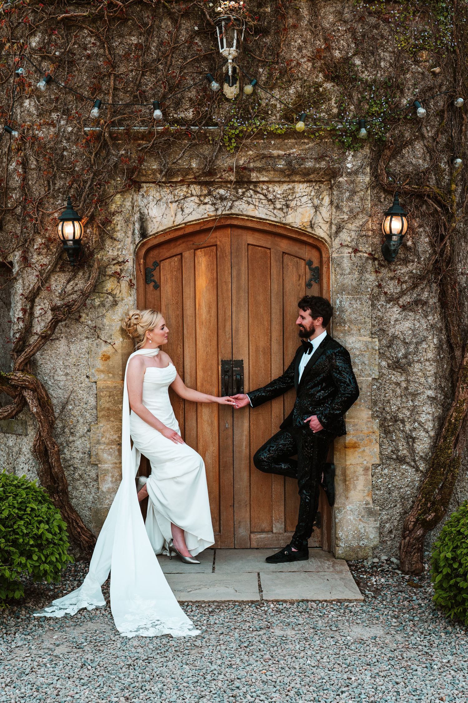 Bride and groom holding hands by a rustic wooden arched door, surrounded by ivy-covered stone walls with vintage lanterns.