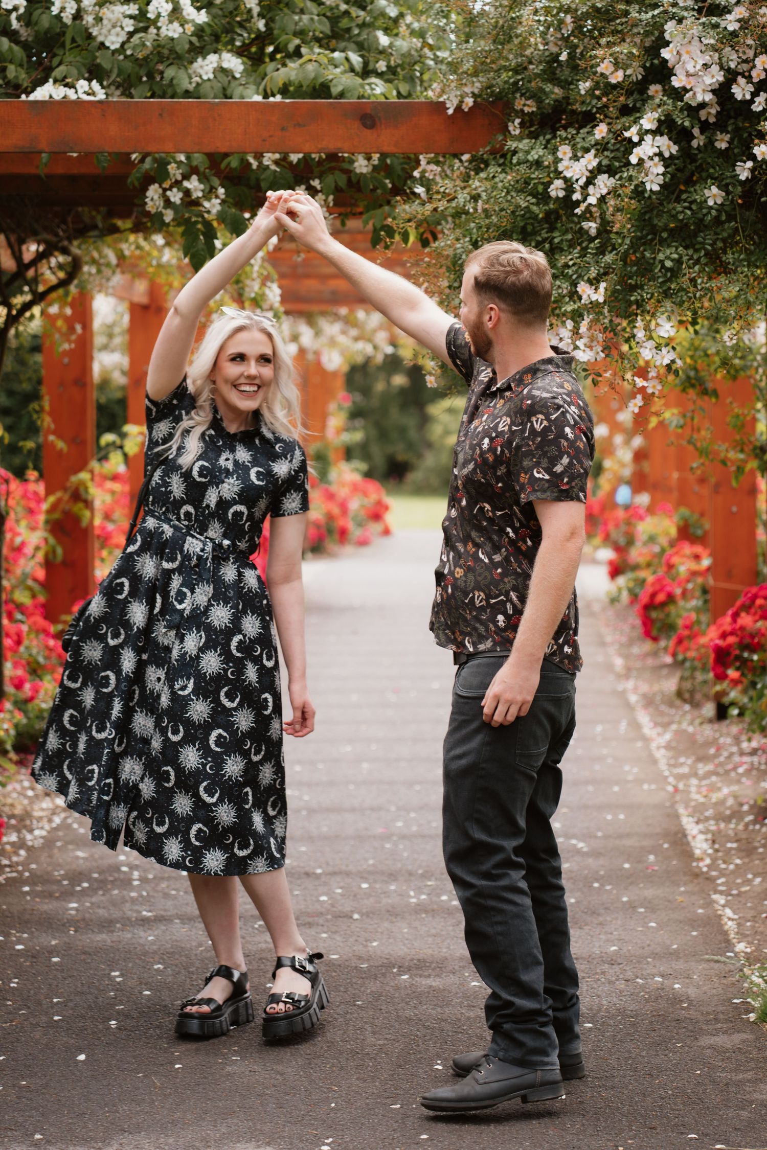 Couple dancing together in a garden pergola with red flowers, woman in black moon dress, man in patterned shirt.