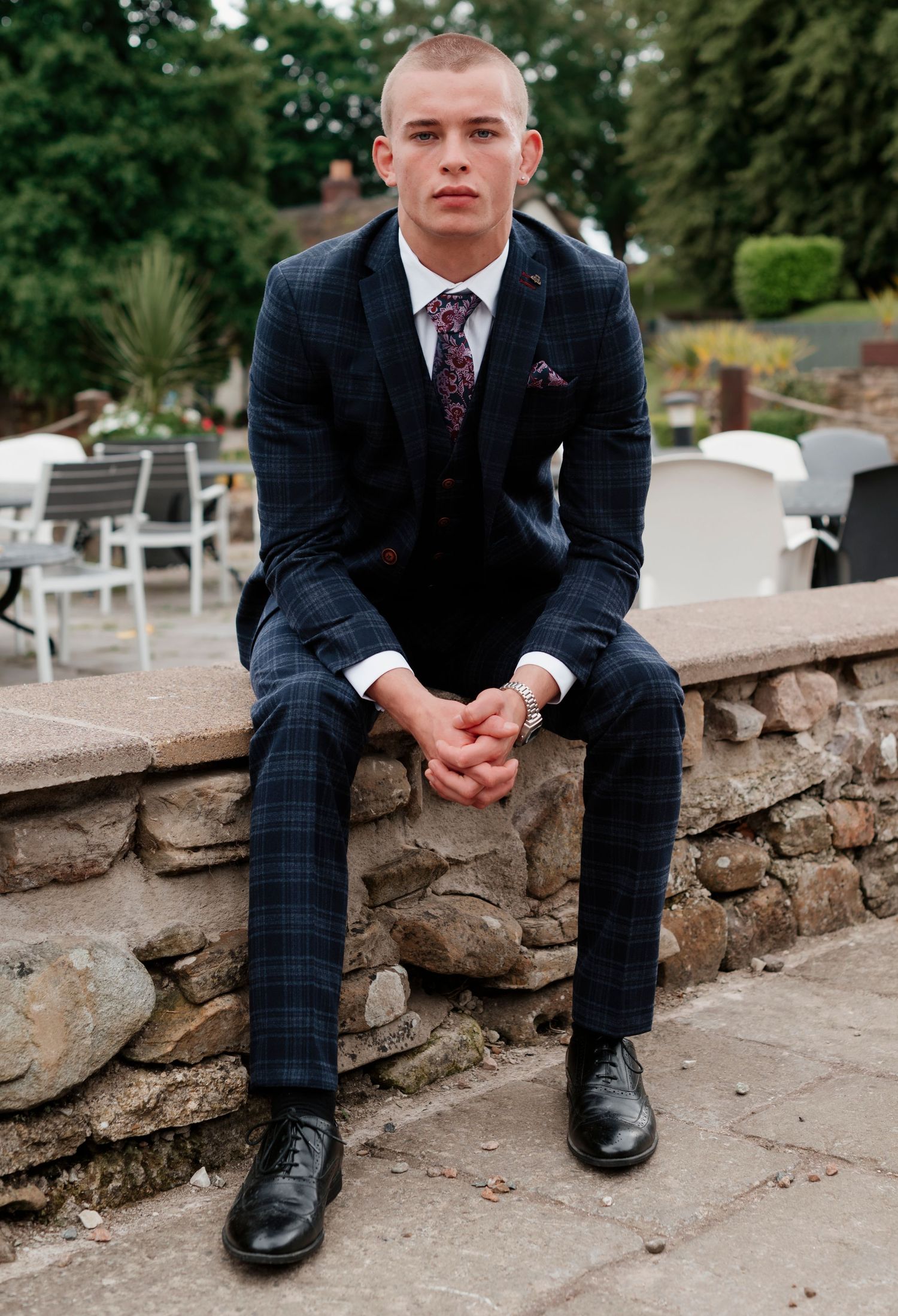Young man in navy plaid suit with tie and pocket square sits on stone wall outdoors at formal event.