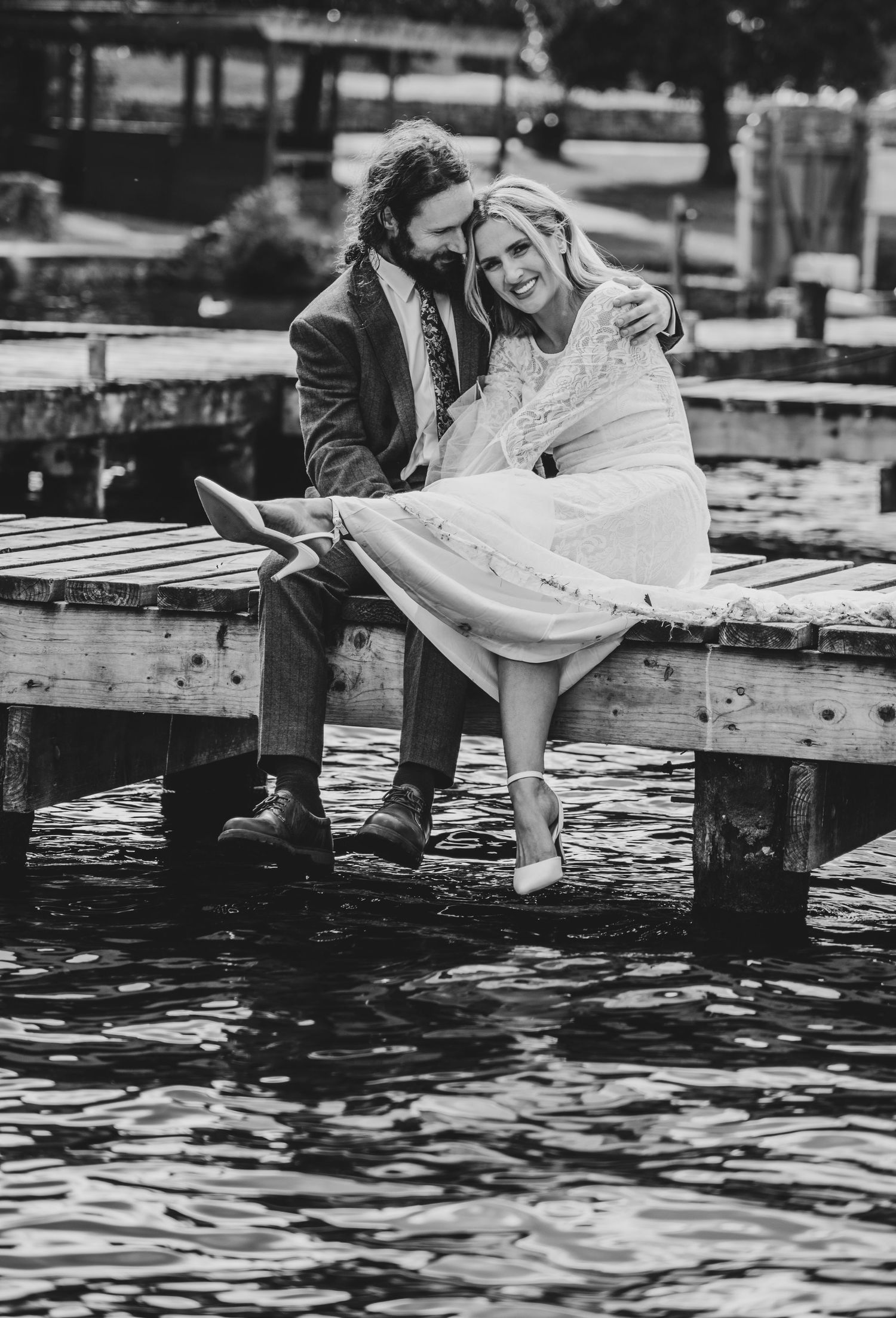 Black and white photo of smiling bride and groom sitting together on wooden dock over water.