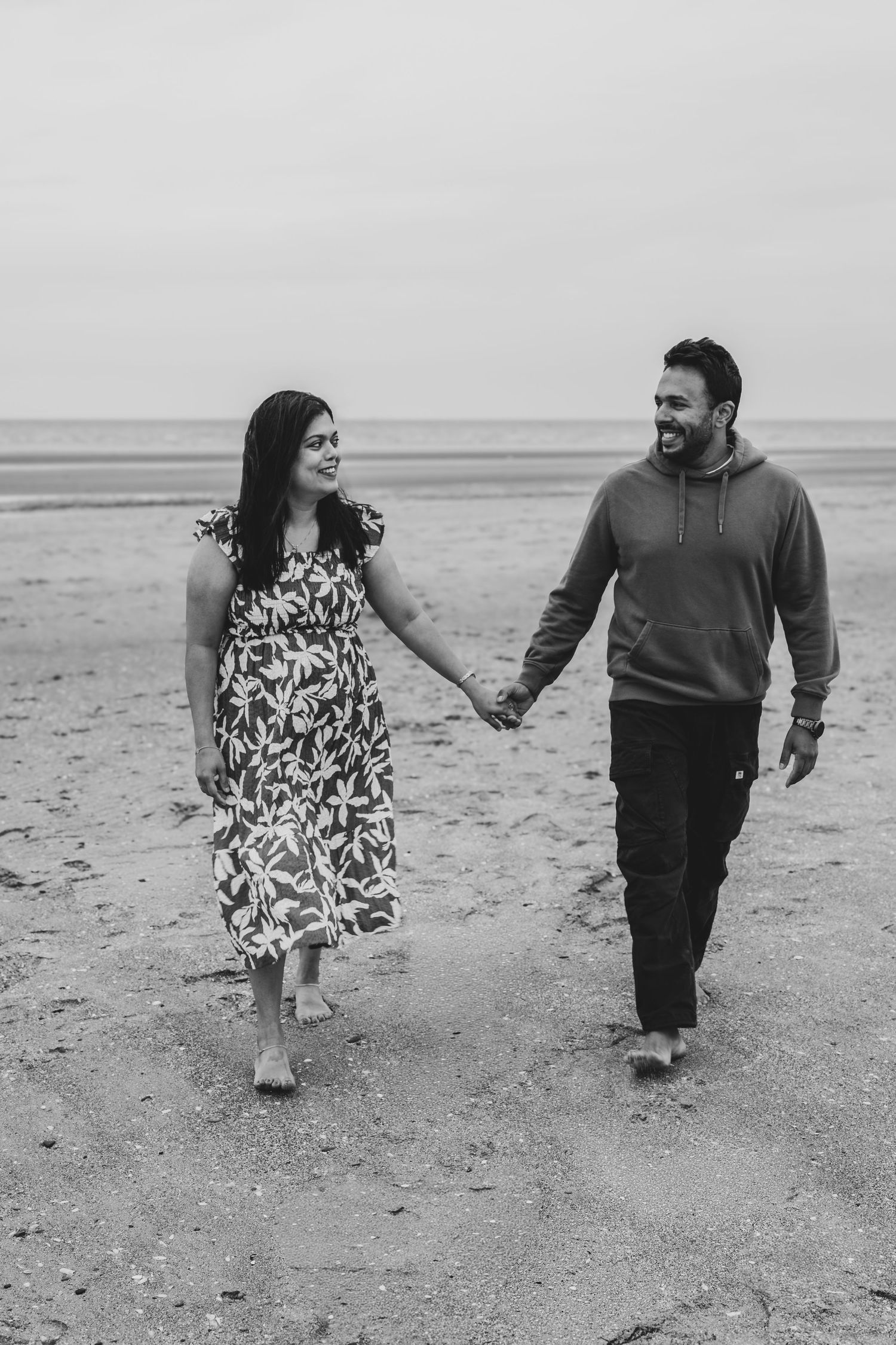 Black and white photo of a happy couple holding hands and walking on a sandy beach, smiling at each other.