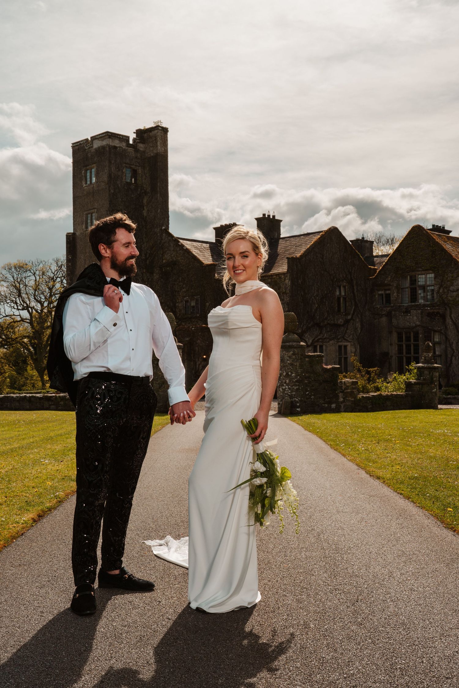 Bride and groom holding hands on driveway of historic castle wedding venue under dramatic cloudy sky.