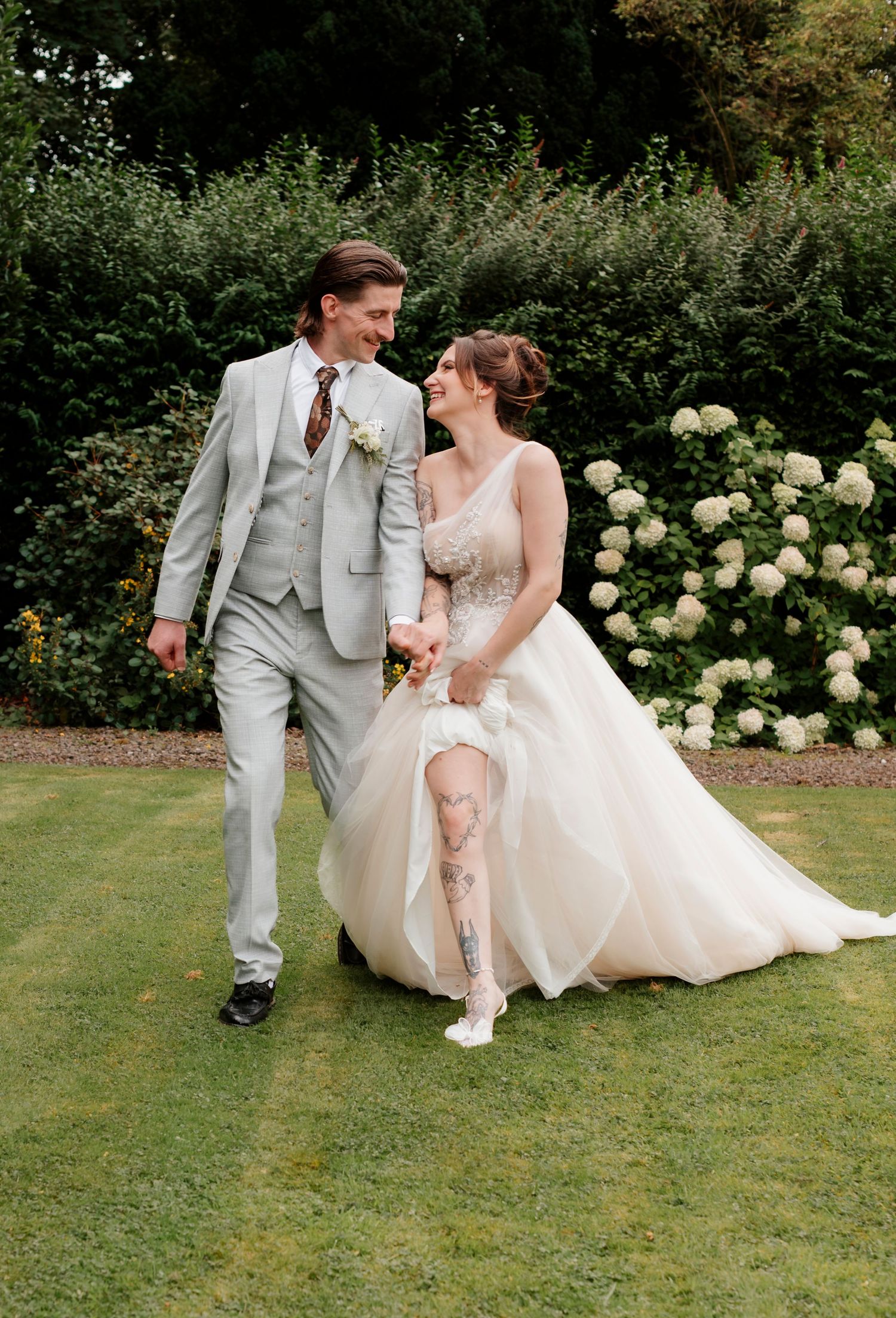 A bride and groom walk hand in hand laughing in a lush garden, she in a ballgown, he in a gray suit.