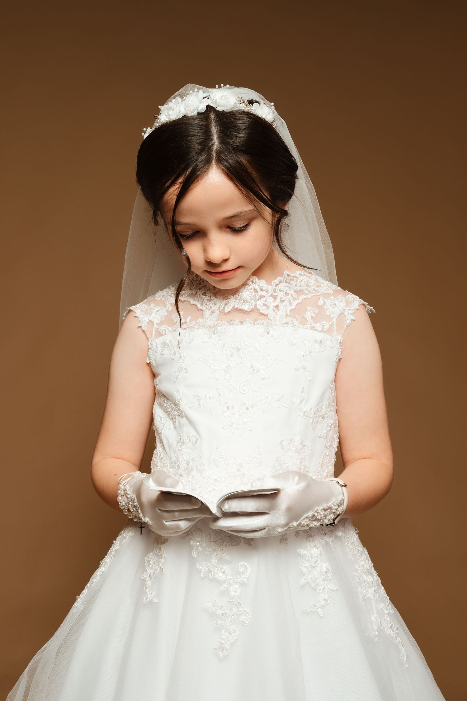 Young girl in white lace First Communion dress, veil, and gloves reading a prayer book on beige background.