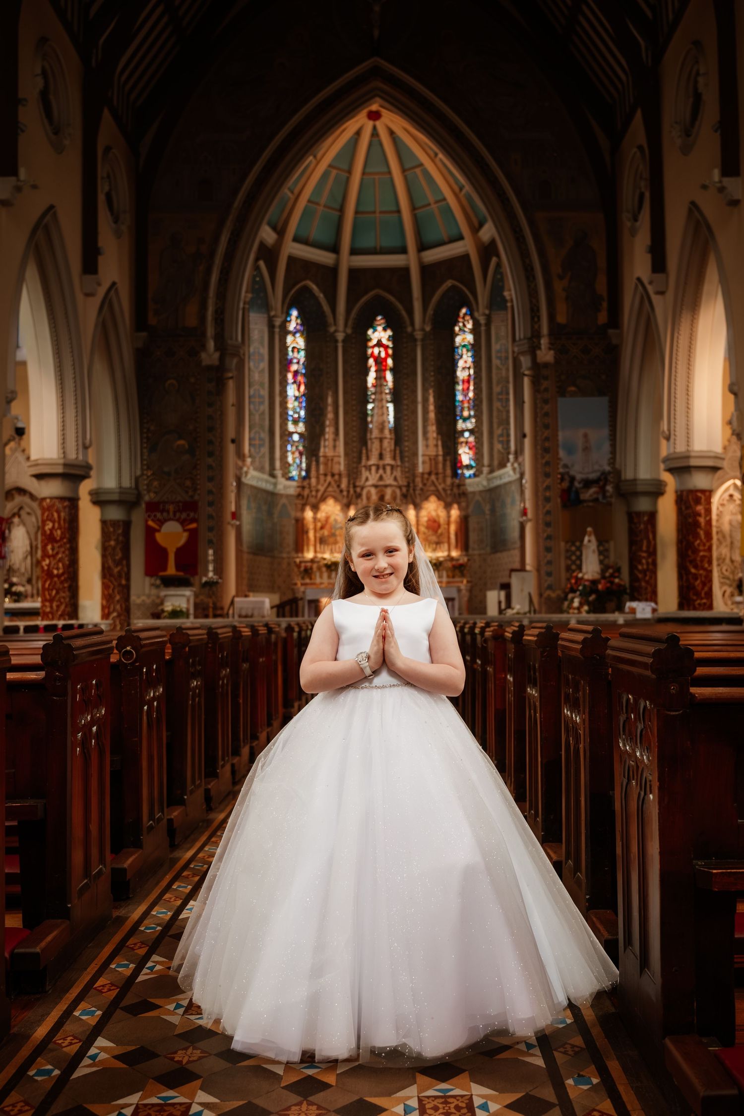 Young girl in white communion dress and veil stands praying in church aisle with stained glass windows.