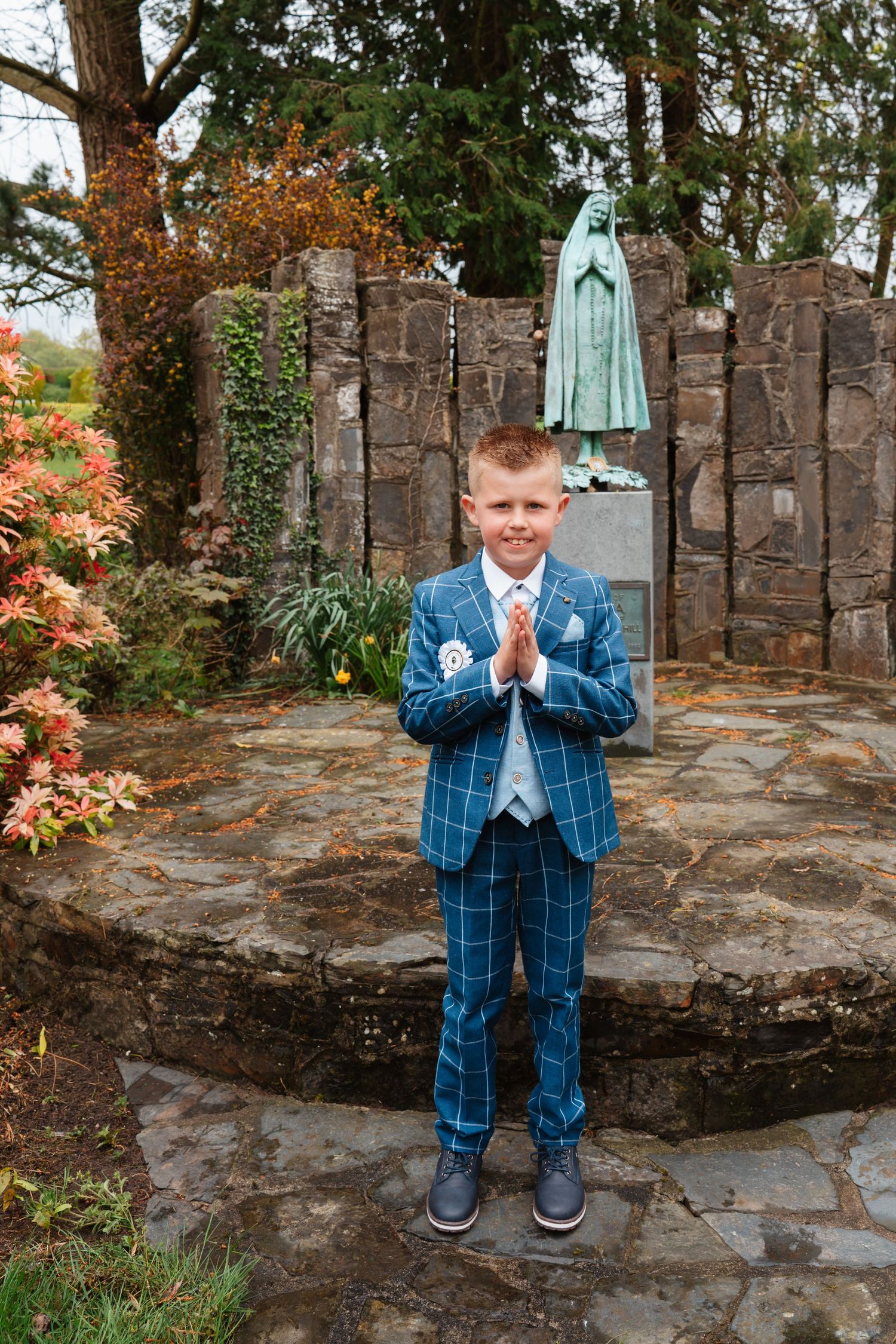 Boy in blue plaid suit with hands clasped in prayer pose standing before a Virgin Mary statue in a garden.
