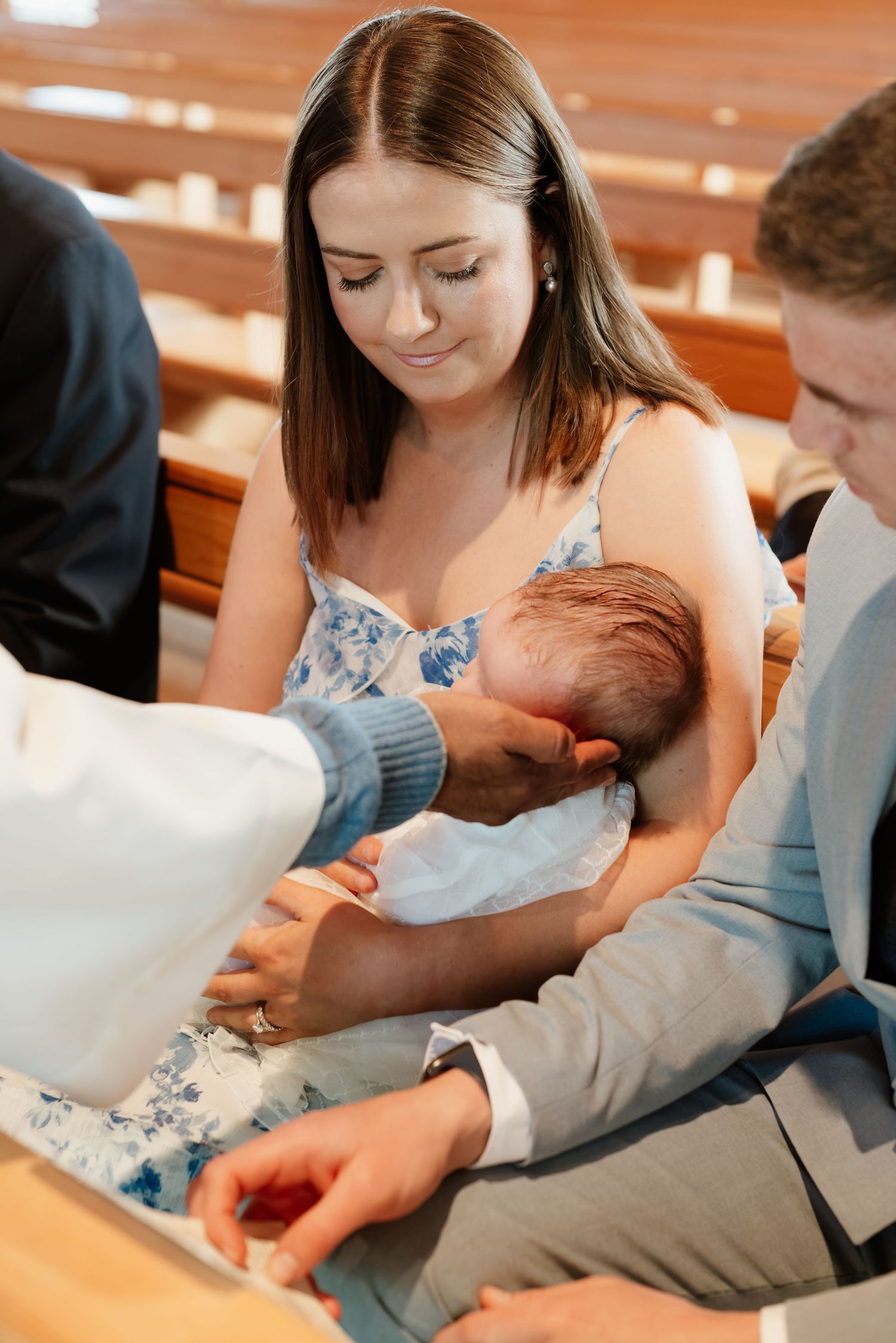 A mother holds her newborn baby during a baptism ceremony in a church, with family members gathered around.