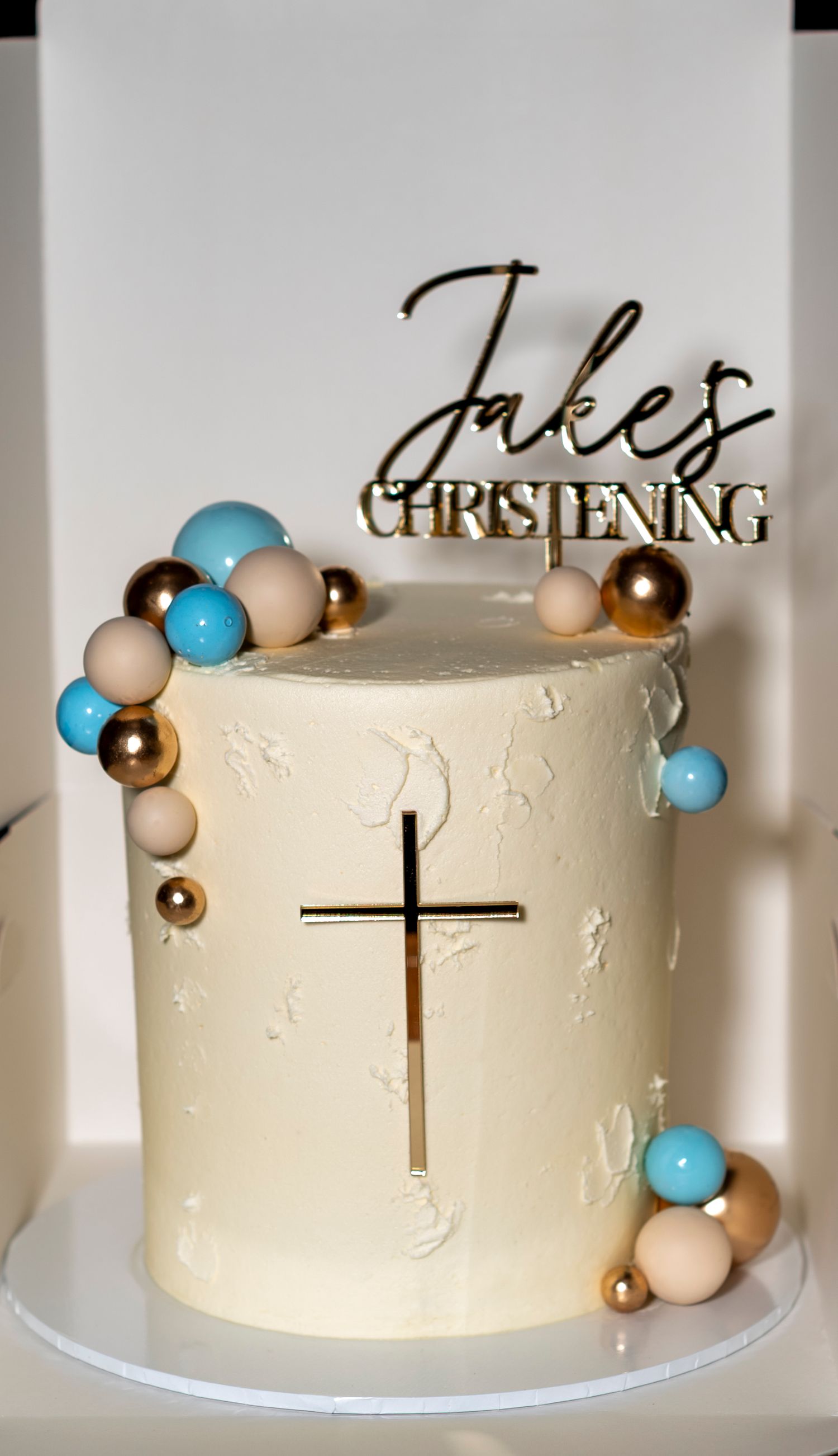 Elegant white christening cake with gold cross and colorful spheres, topped with Jakes Christening topper.