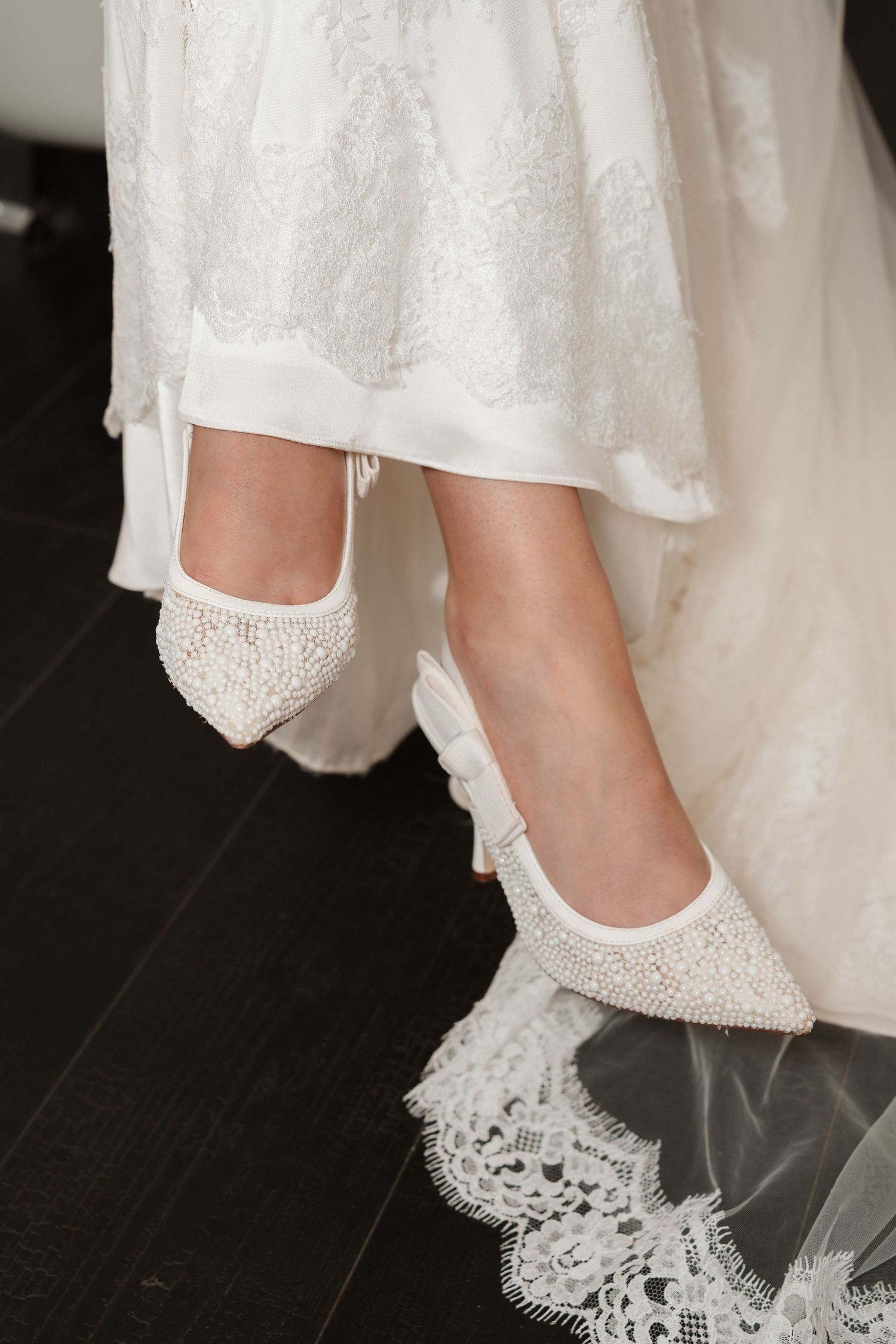 Close-up of bride wearing elegant pearl-embellished pointed-toe white wedding heels with lace bridal gown.