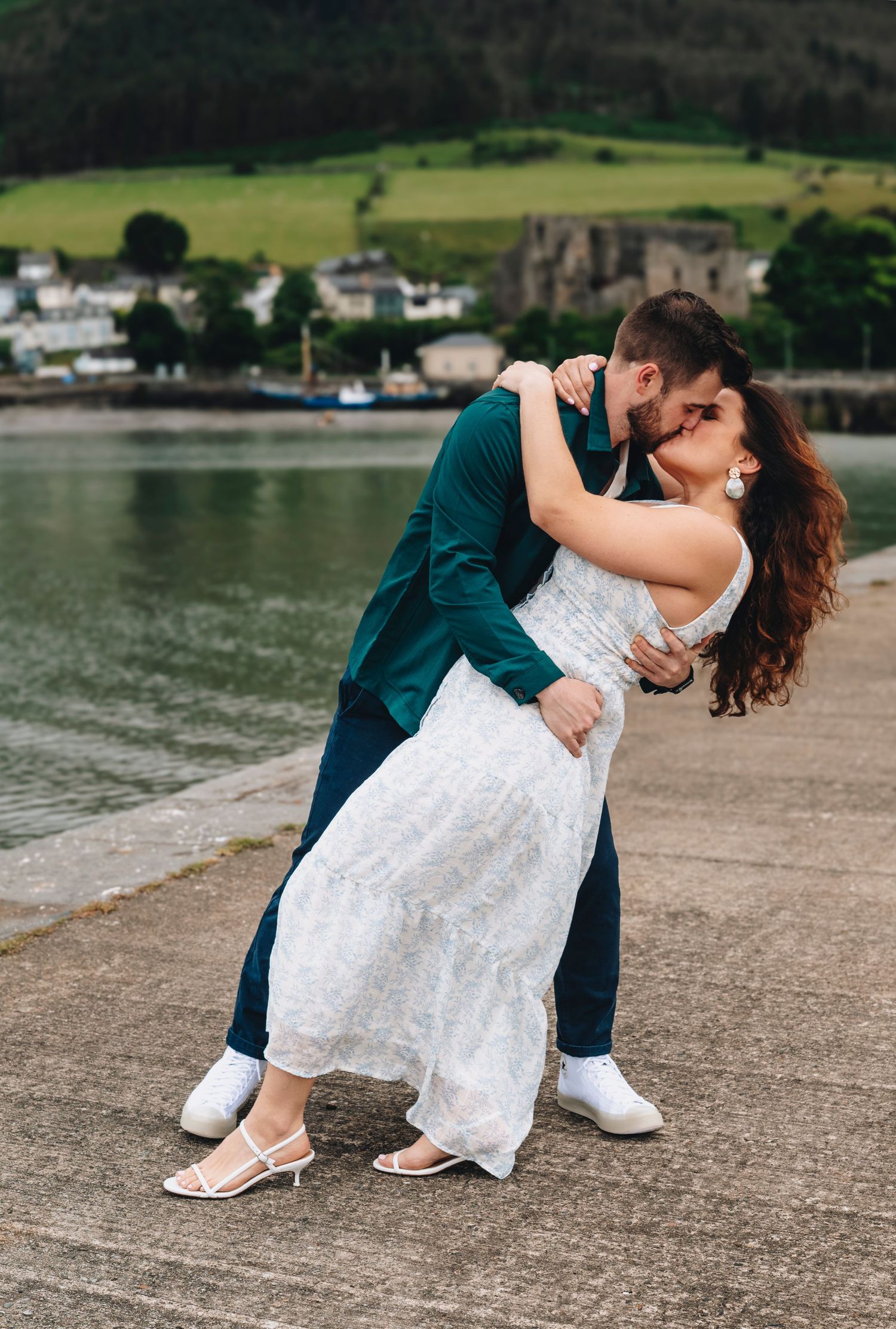 Couple sharing a romantic dip kiss on a waterfront pier, bride in white lace dress and groom in teal suit.