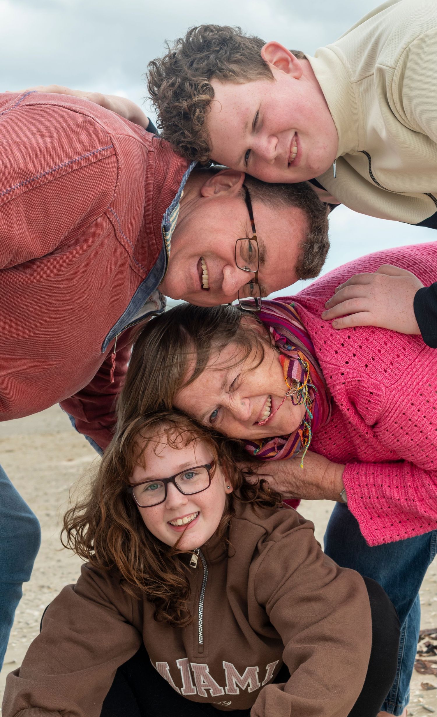 Family of four joyfully stacking heads outdoors on a beach, smiling and laughing together on a cloudy day.