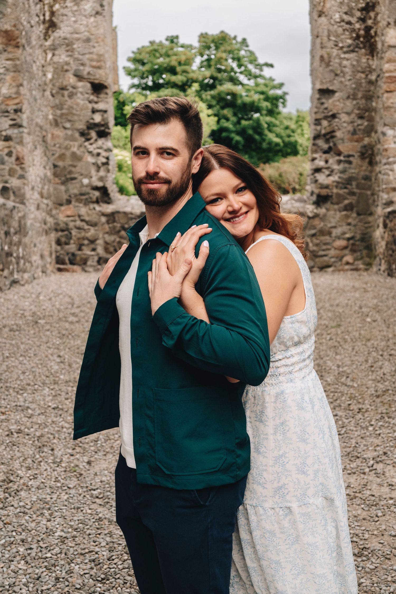 Couple posing at stone ruins, woman in white dress hugging man in teal jacket, showing engagement ring.