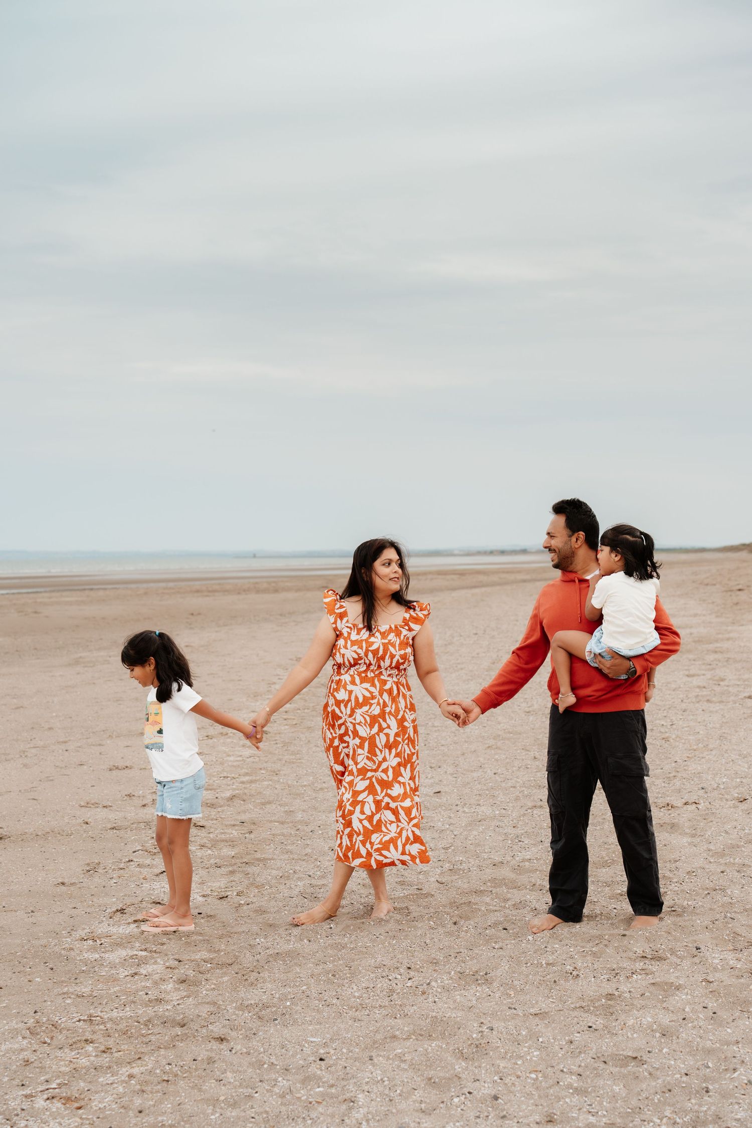 A happy family of four holds hands on a sandy beach, with parents in red outfits and two young daughters.