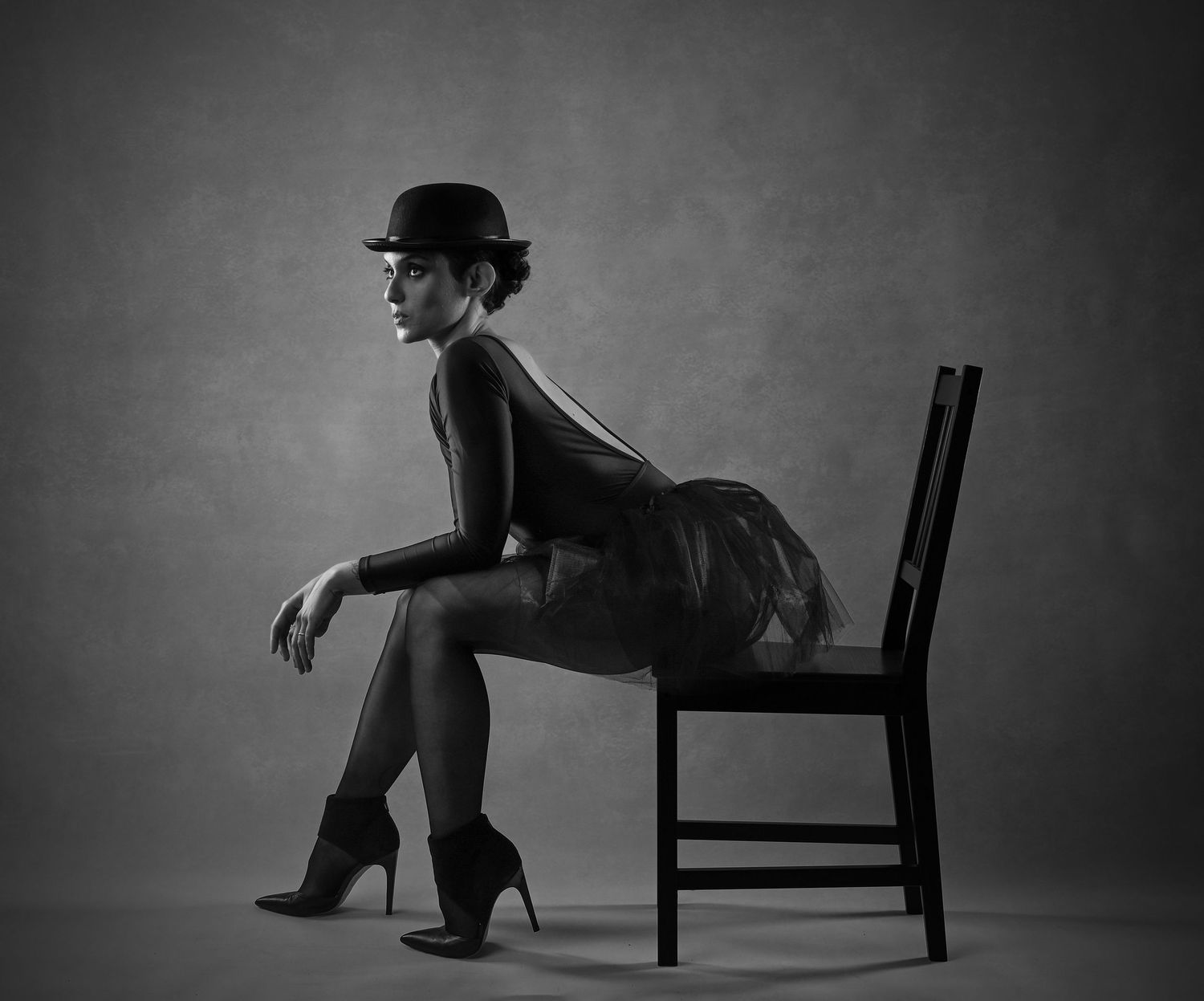 Black and white photo of a woman in a bowler hat, tutu skirt, and heels sitting elegantly on a wooden chair.
