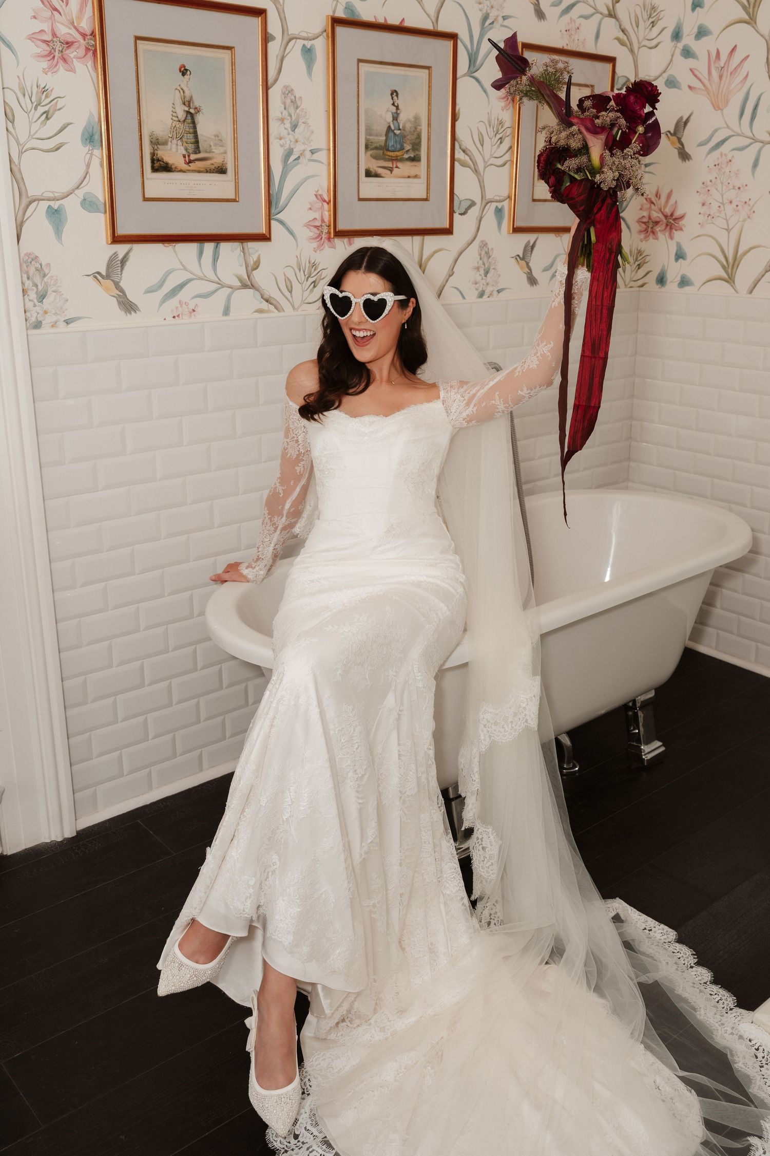 Bride in white off-shoulder gown, heart sunglasses, and veil poses by clawfoot tub in floral wallpaper bathroom.
