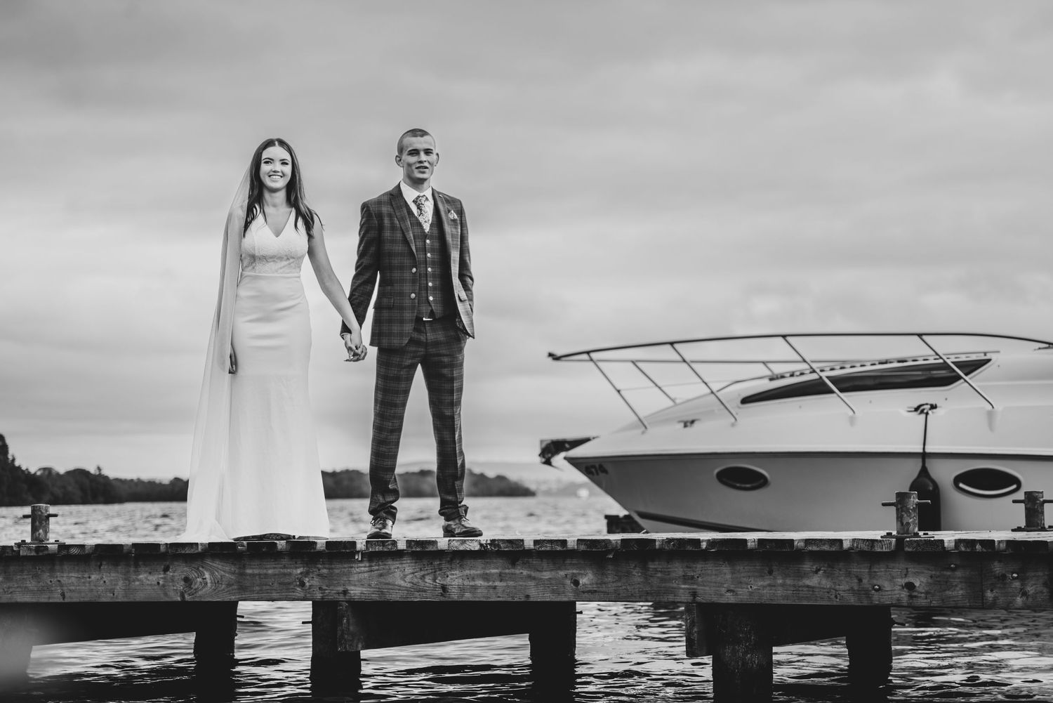 Black and white photo of bride and groom standing on wooden dock with boat on cloudy day.