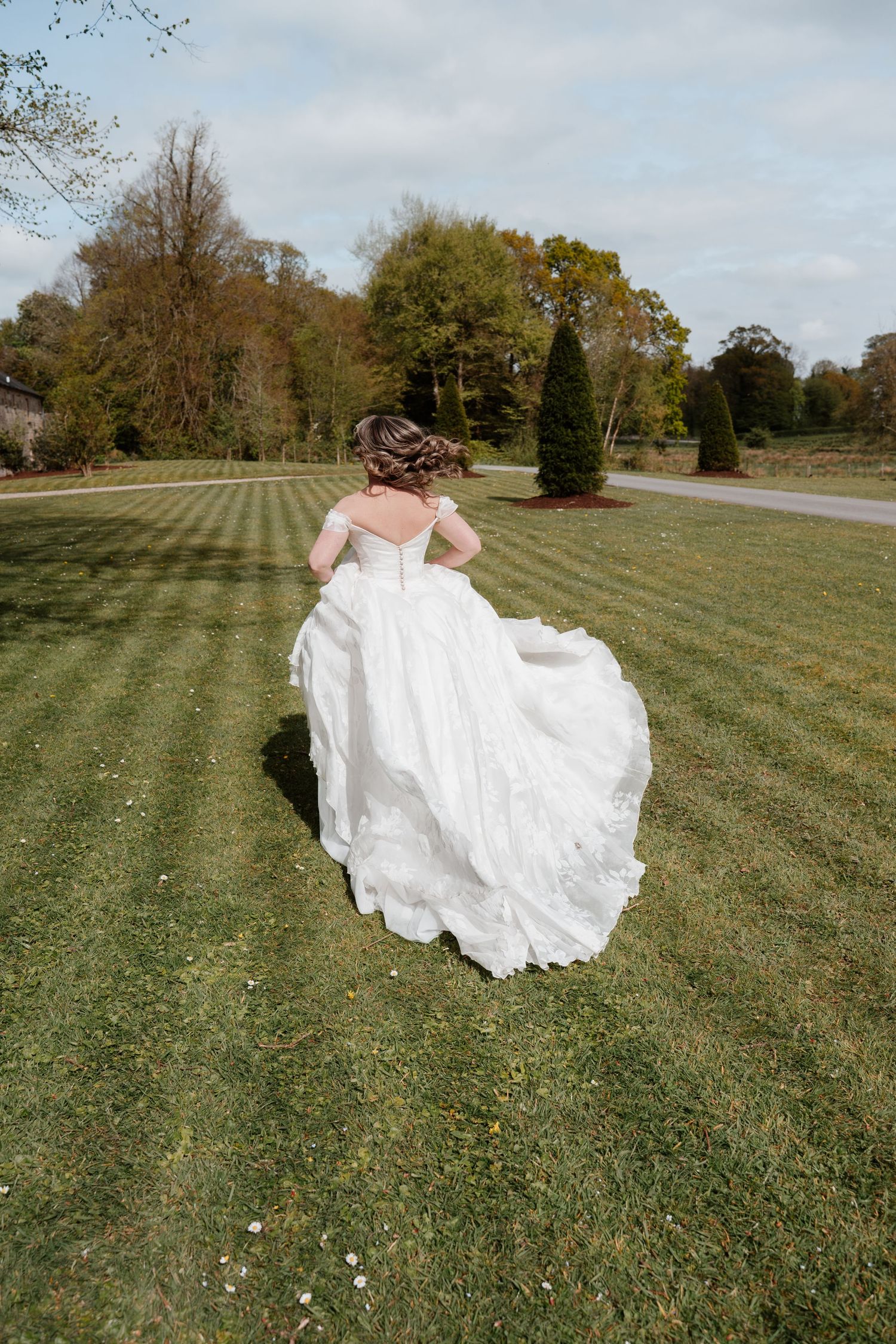 Bride in a flowing white ballgown walking across a manicured lawn with trees in the background.