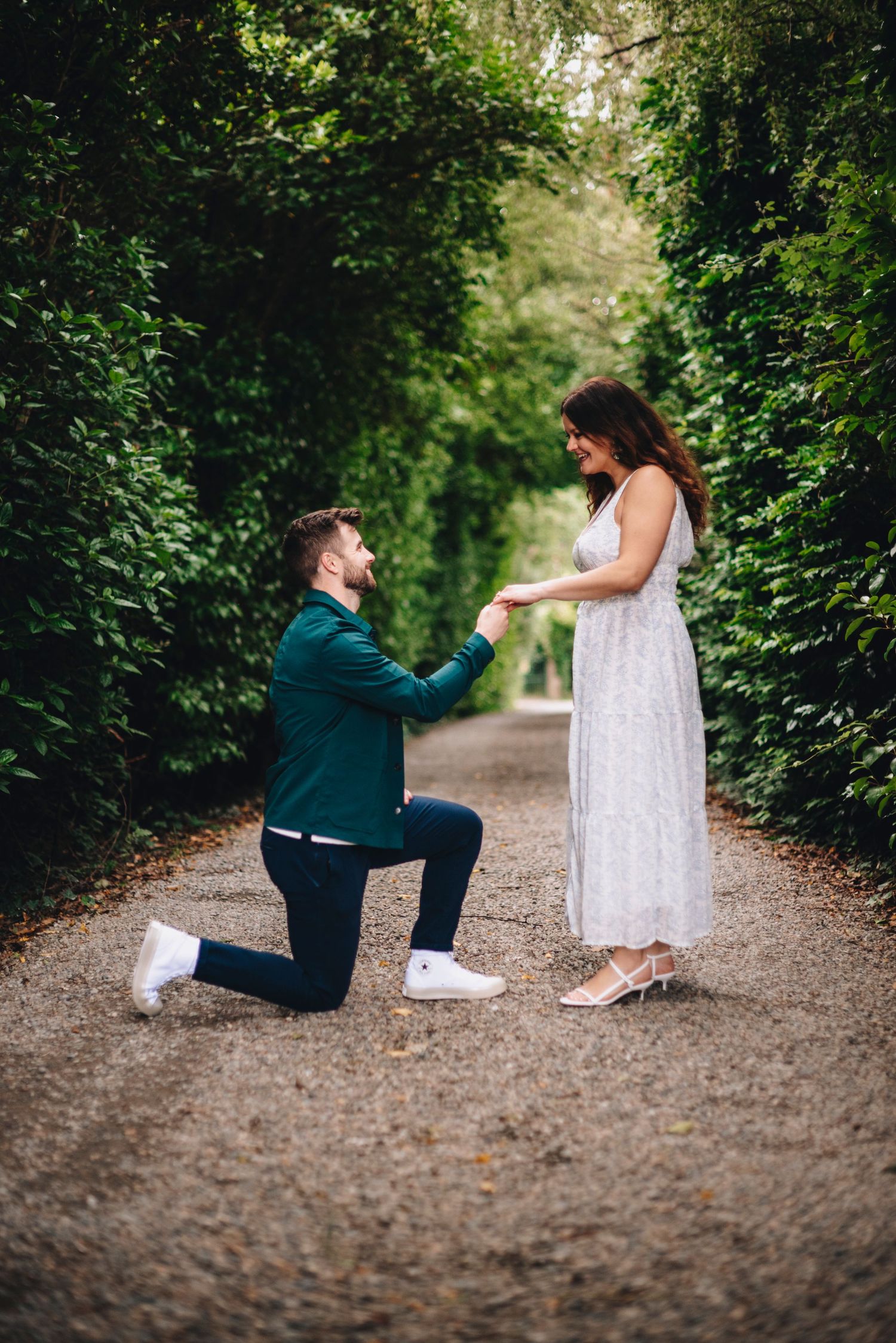 Man on one knee proposing to woman in white dress on a tree-lined gravel path during an outdoor engagement.