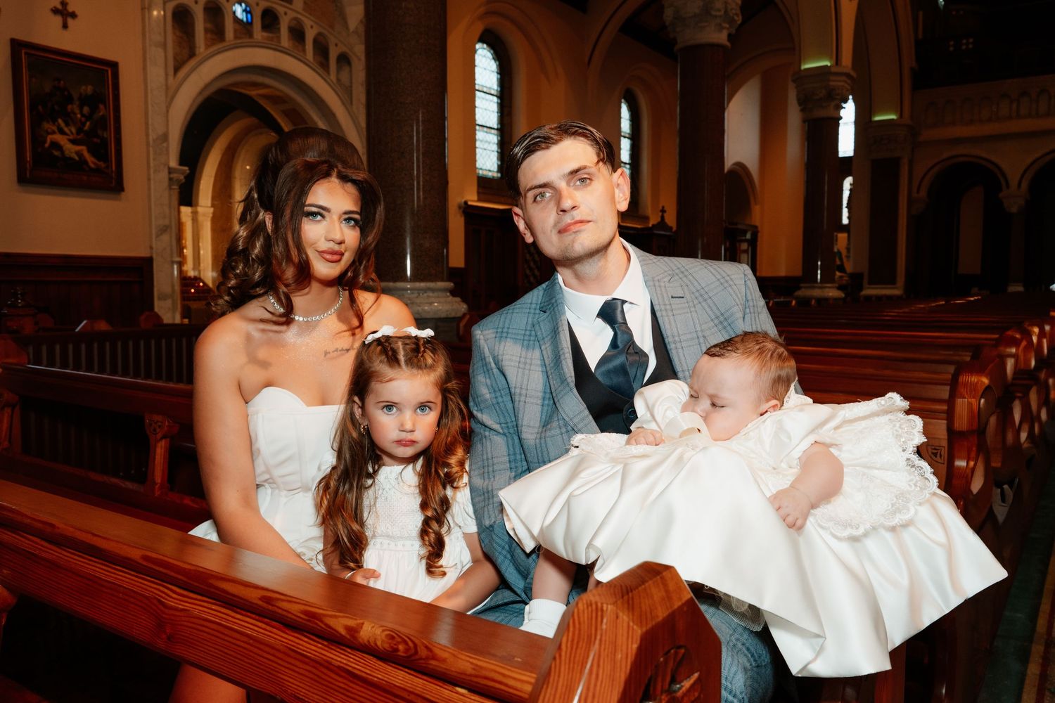 Family posing in church pew during baptism ceremony, parents with young girl and baby in white christening gown.