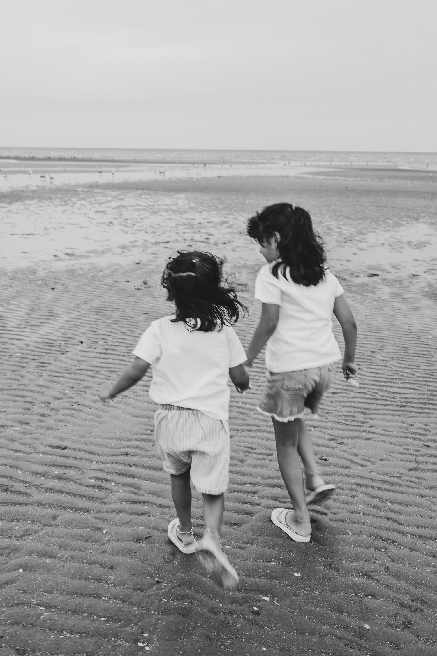 Two children holding hands and running on a sandy beach in black and white photography.