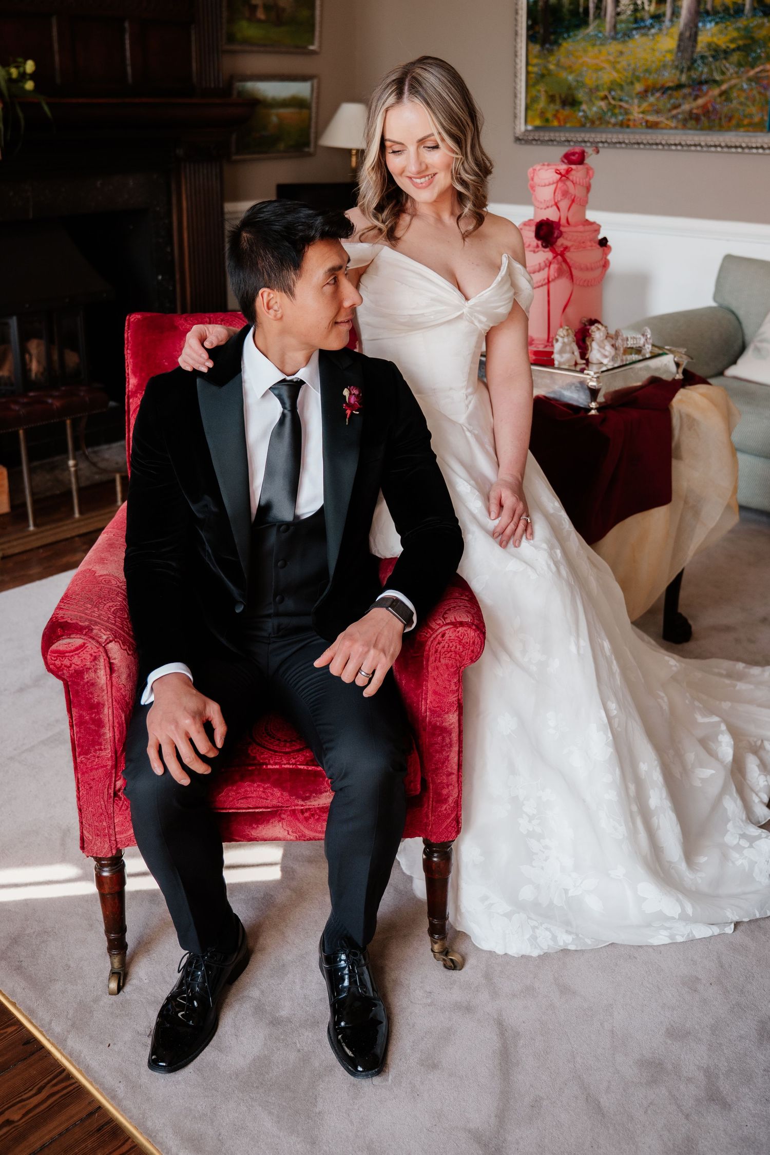Groom in black tuxedo sits in red velvet chair as smiling bride in off-shoulder gown stands beside him.