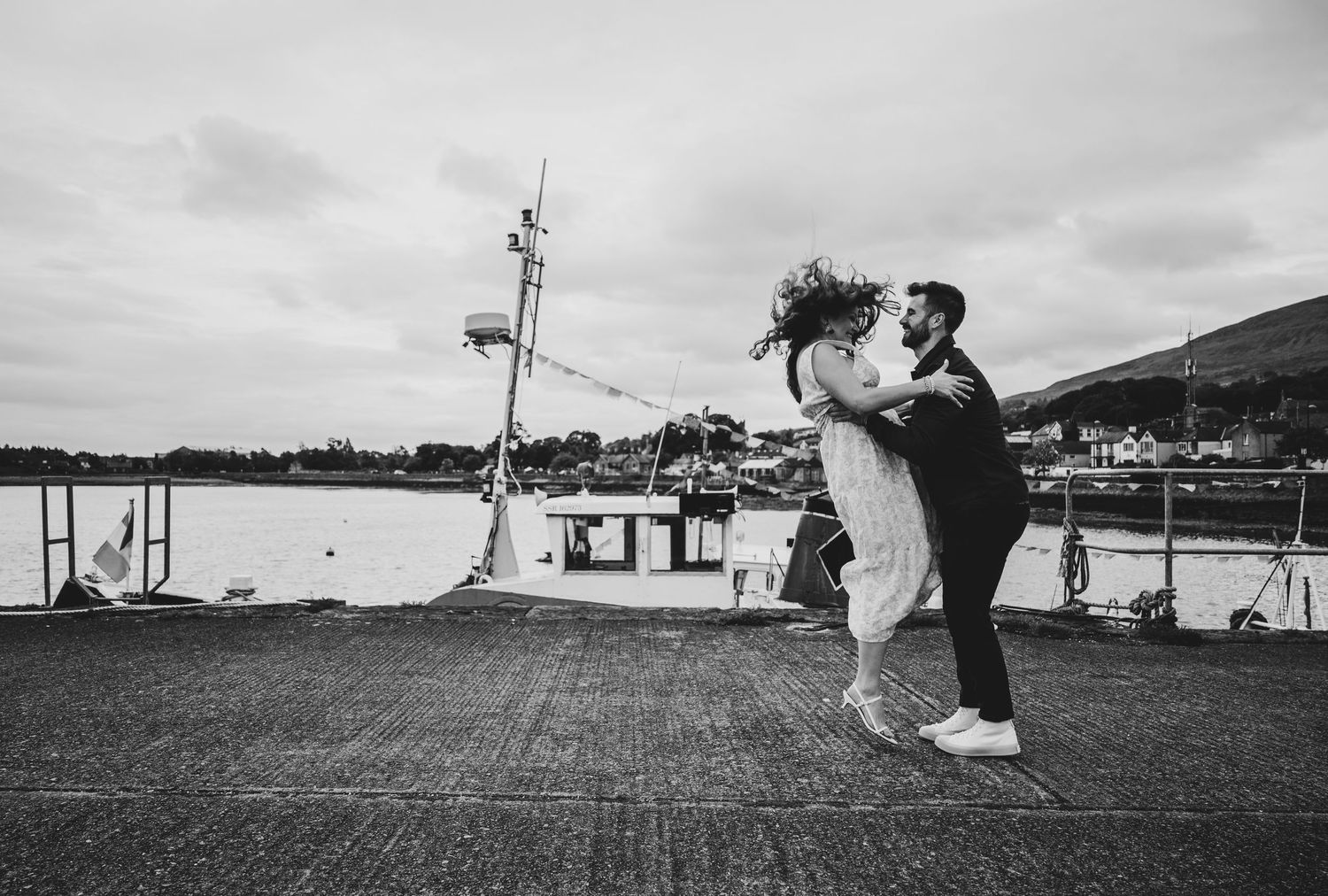 Black and white photo of groom lifting bride on a harbor pier waterfront.